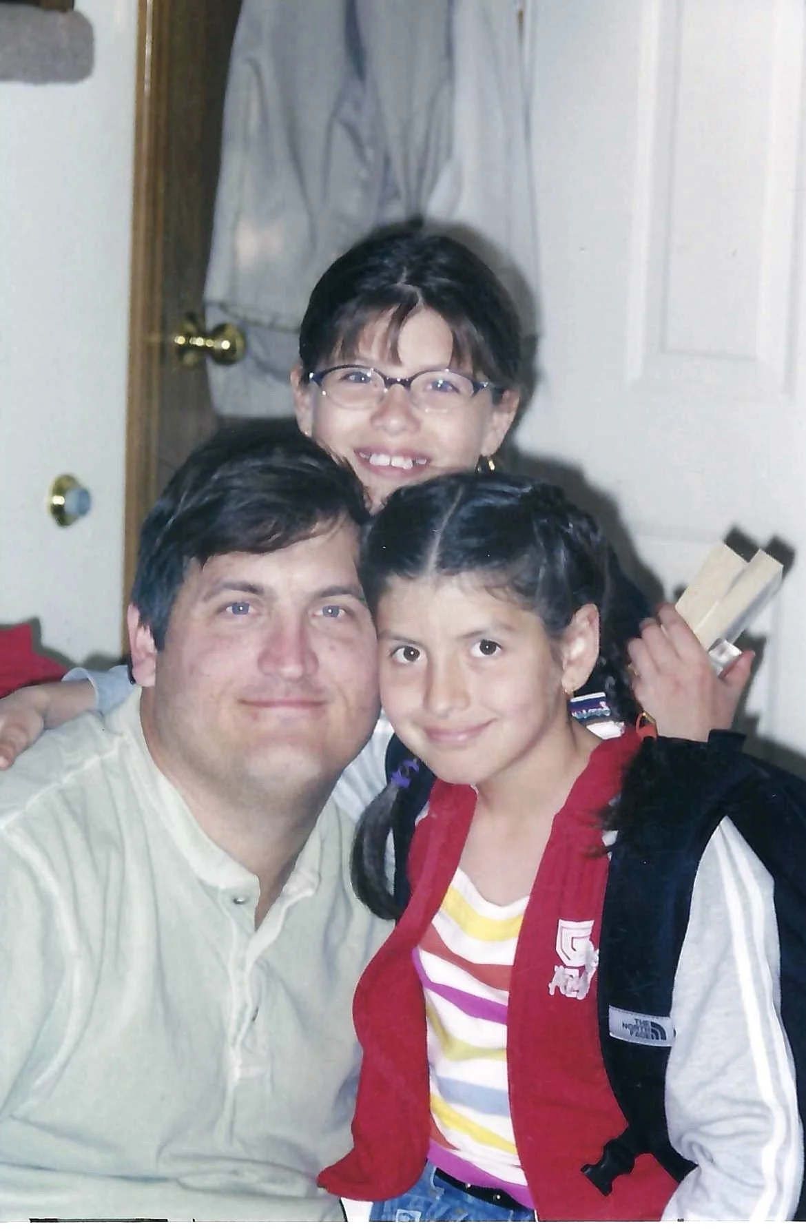 A group of three people, two adults and a girl, smiling and posing indoors. The girl has braided hair, is wearing a red vest, a striped shirt, and has a backpack. The man has dark hair, is wearing a light-colored shirt, and the woman has dark hair, g