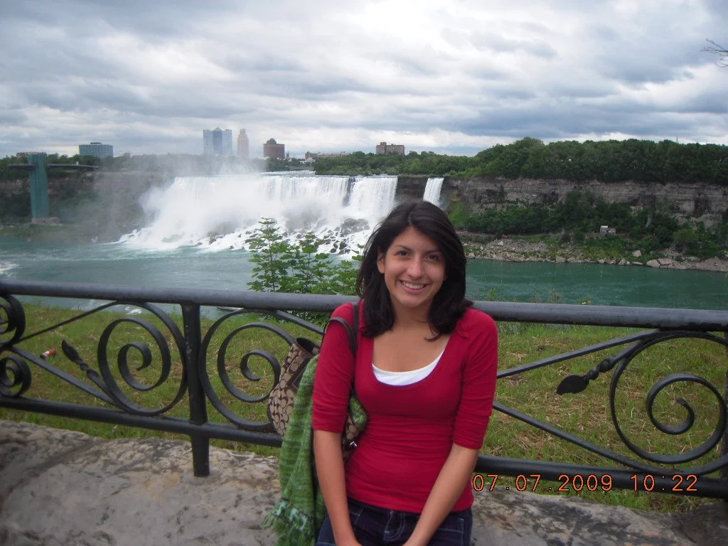 A young woman smiling in front of Niagara Falls, with a city skyline and cloudy sky in the background.