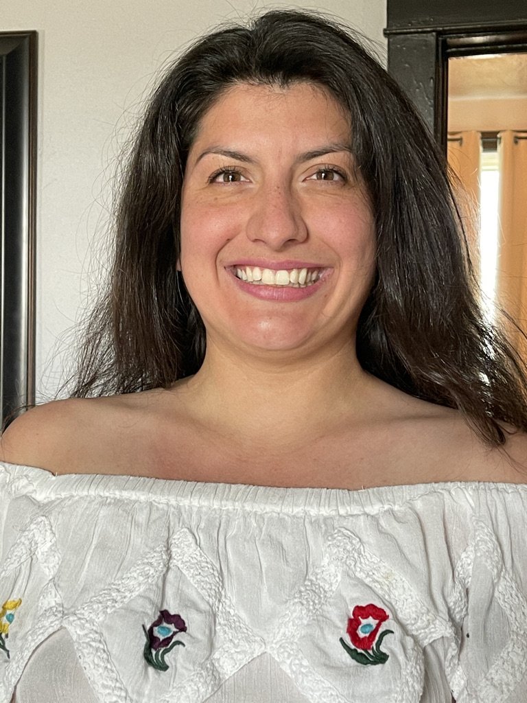 A woman with long dark hair smiling at the camera, wearing an off-the-shoulder white top with embroidered floral designs.