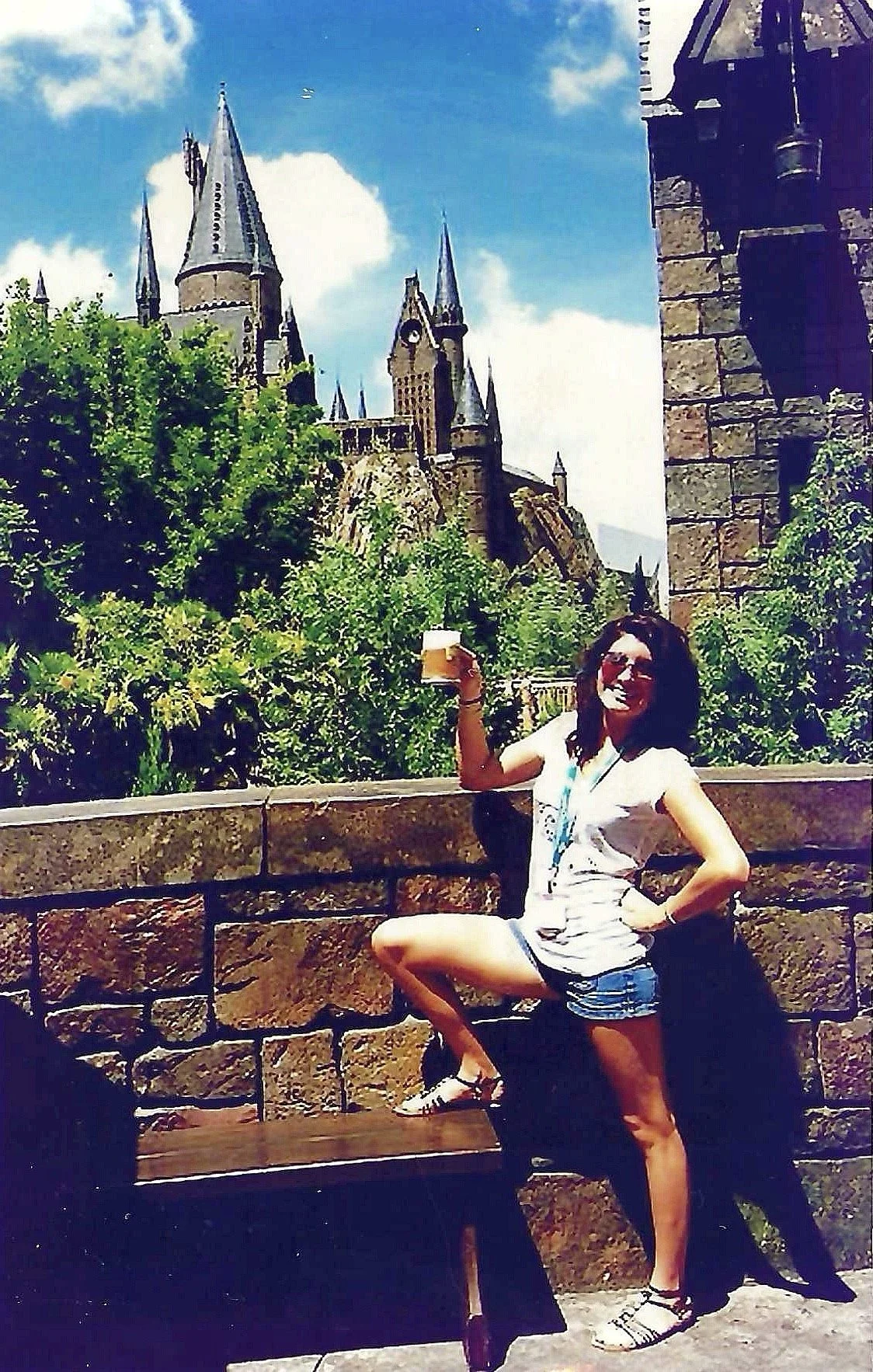 A woman dressed in a white tank top and shorts, holding a drink, posing playfully in front of a castle with tall spires, green bushes, and blue sky with clouds.
