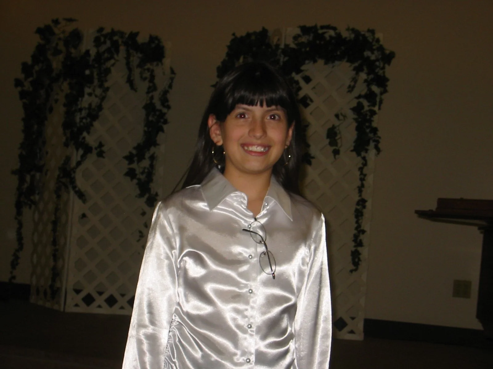 A woman with dark hair wearing a shiny, cream-colored satin shirt, smiling indoors with decorative ivy and lattice in the background.