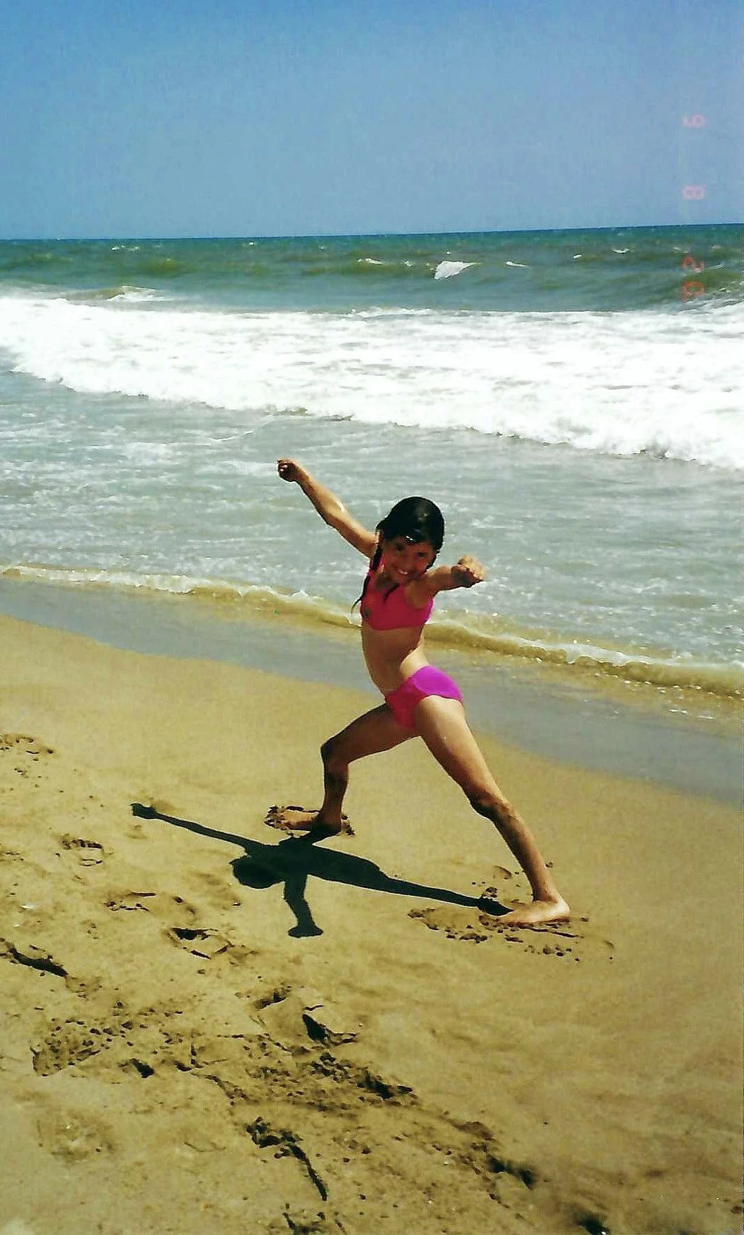 A young girl in a pink swimsuit playing and striking a martial arts pose on the sandy beach near the ocean.