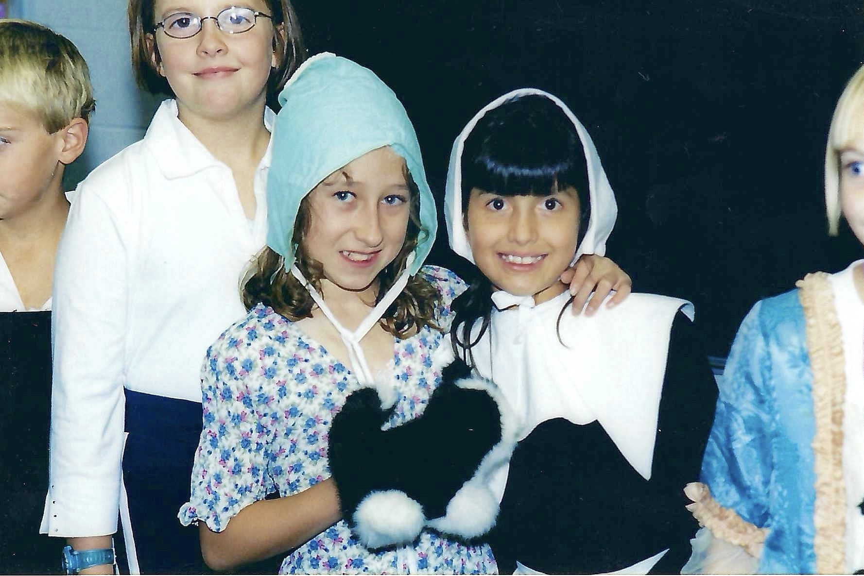 Group of children dressed in historical costumes, including bonnets, dresses, and vests, standing together in front of a black background.