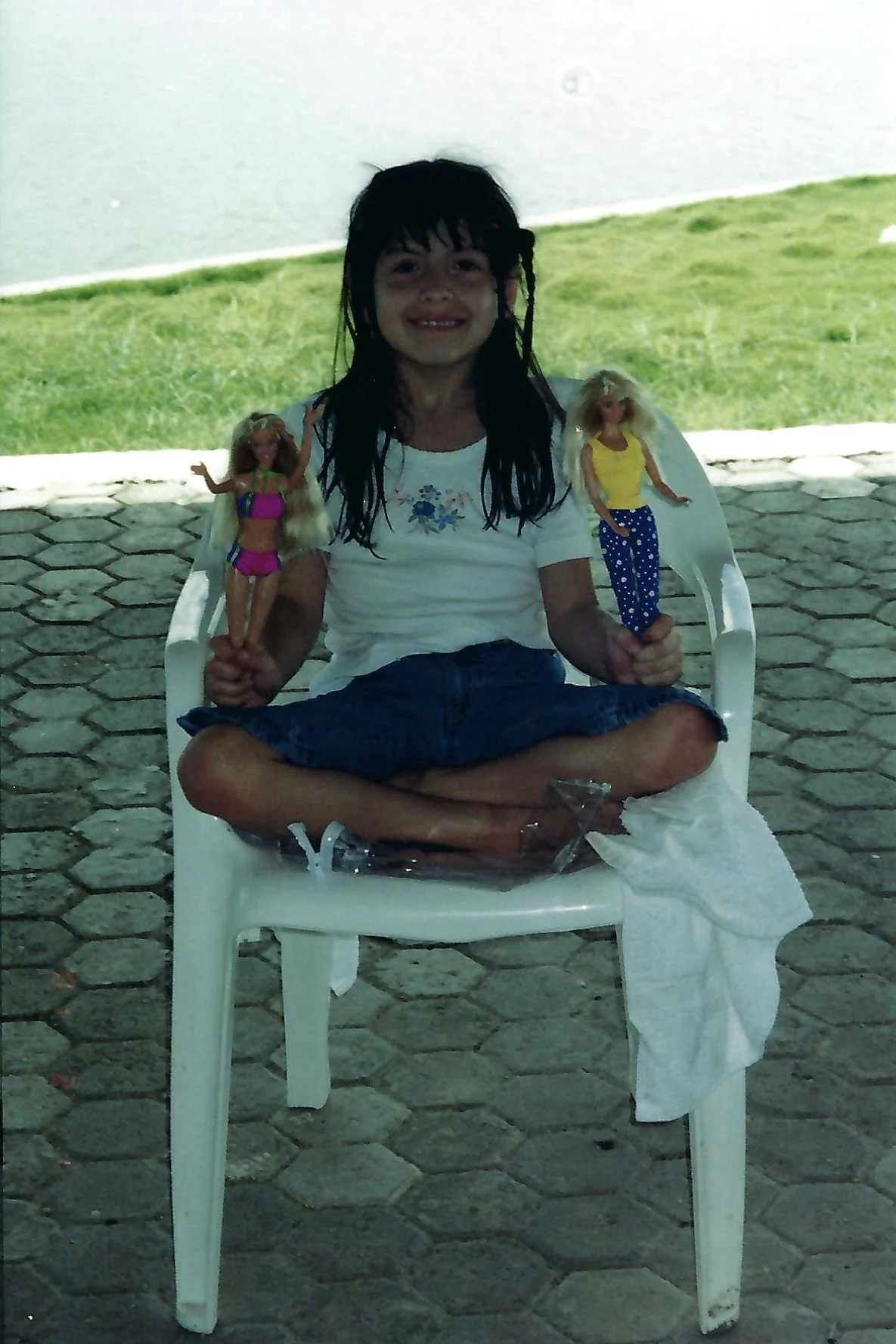Young girl sitting on a white plastic chair outdoors, holding two Barbie dolls, with a grassy background.