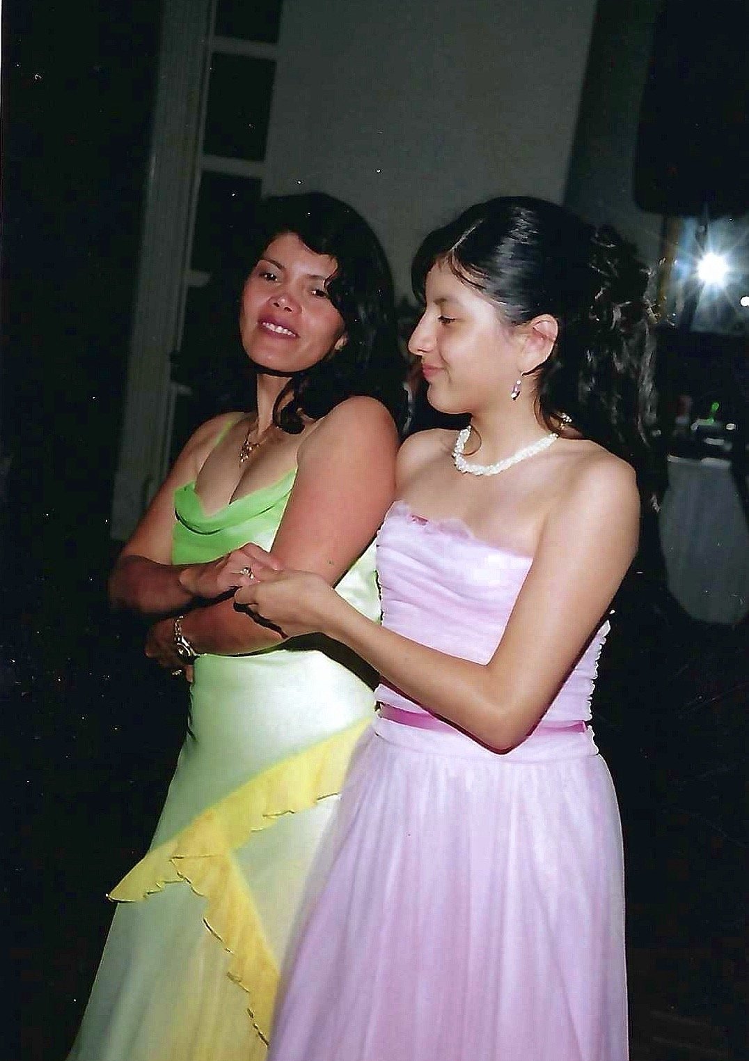 Two women in formal dresses, one in a yellow and green gown and the other in a pink gown, standing close together at a celebration.