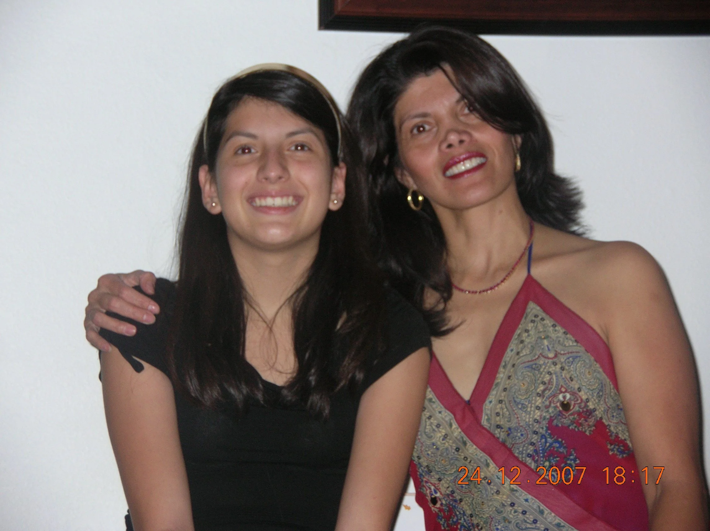 Two women smiling, one with light skin and dark hair wearing a black top, and the other with darker skin and dark hair in a colorful dress, standing close together against a plain white wall.