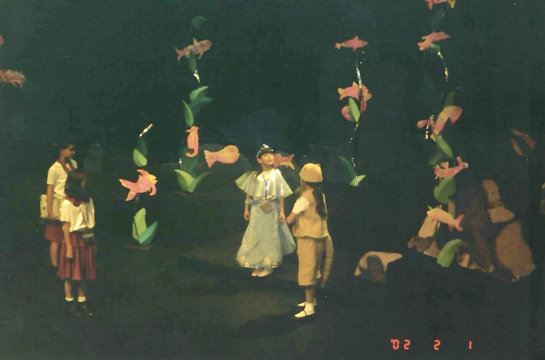 Children dressed in costumes standing on stage with fish and plant decorations, appearing to perform a play or skit.