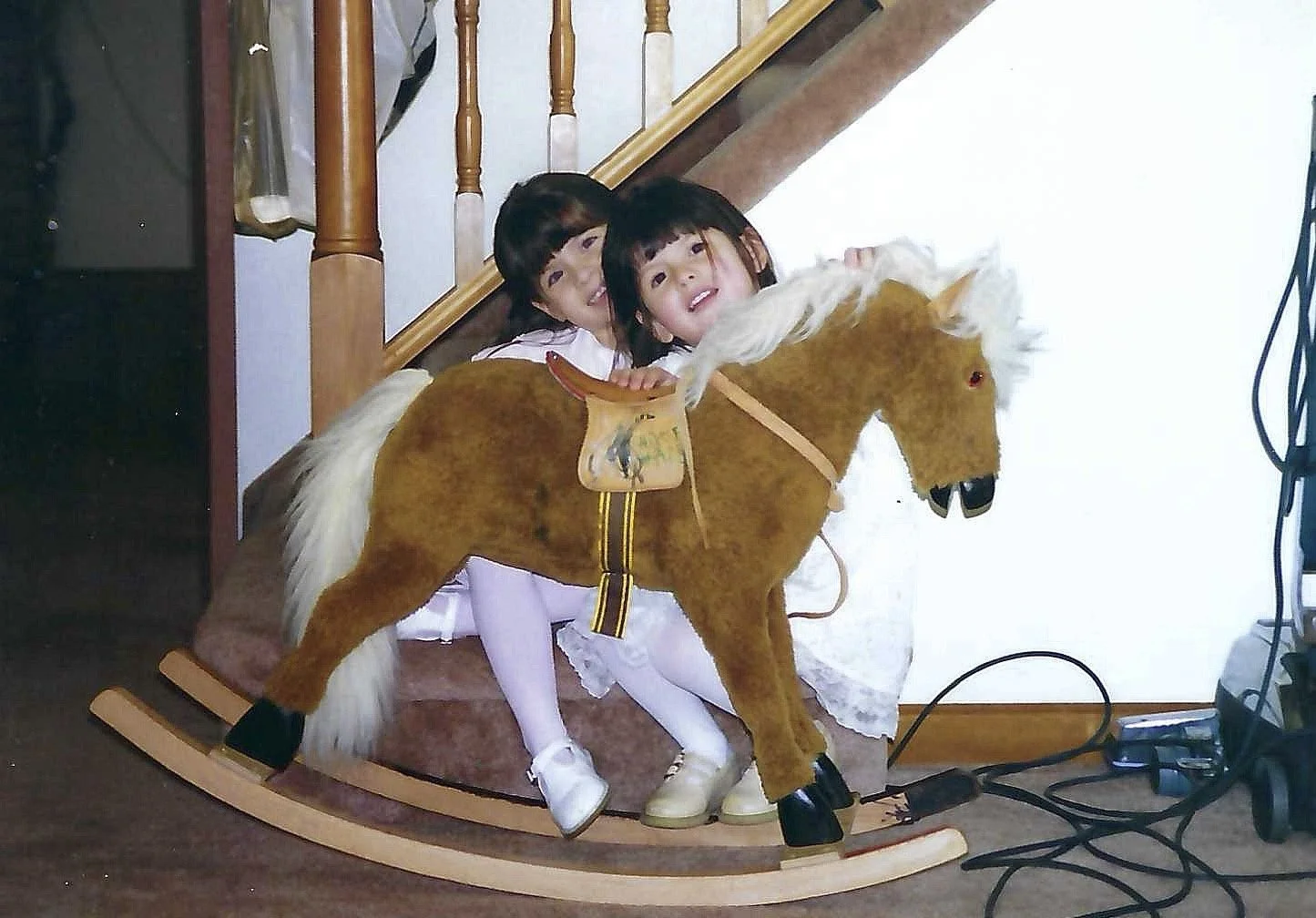 Two young children sitting on a rocking horse in a room near a staircase, with visible electrical cords on the floor.