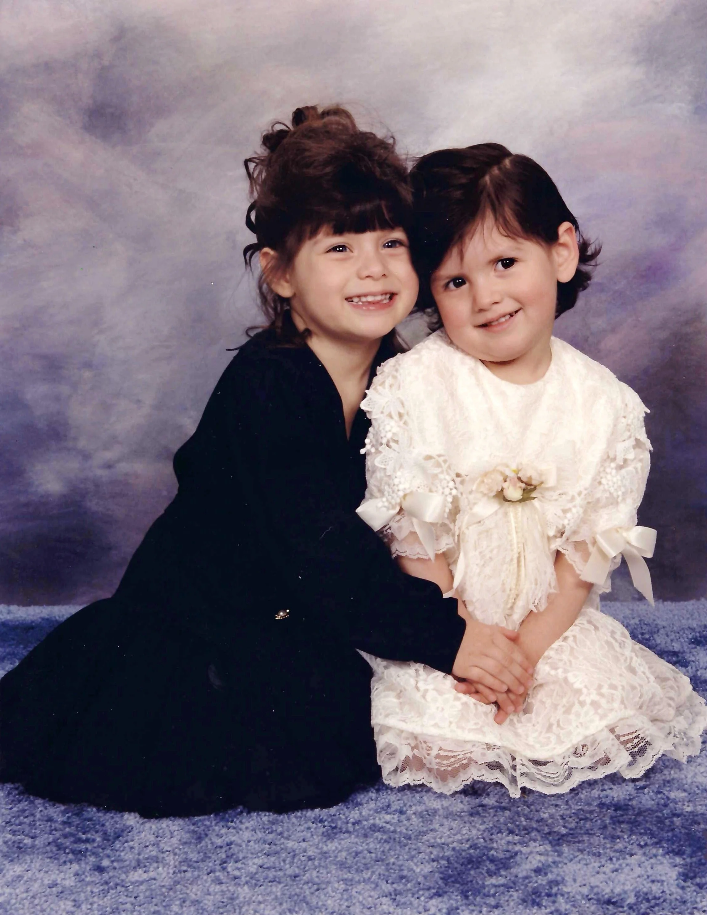 A woman with curly brown hair and a young girl with short dark hair, both smiling and sitting together on a textured blue surface against a cloudy, purplish background. The girl is wearing a cream-colored lace dress with bows, and the woman is wearin
