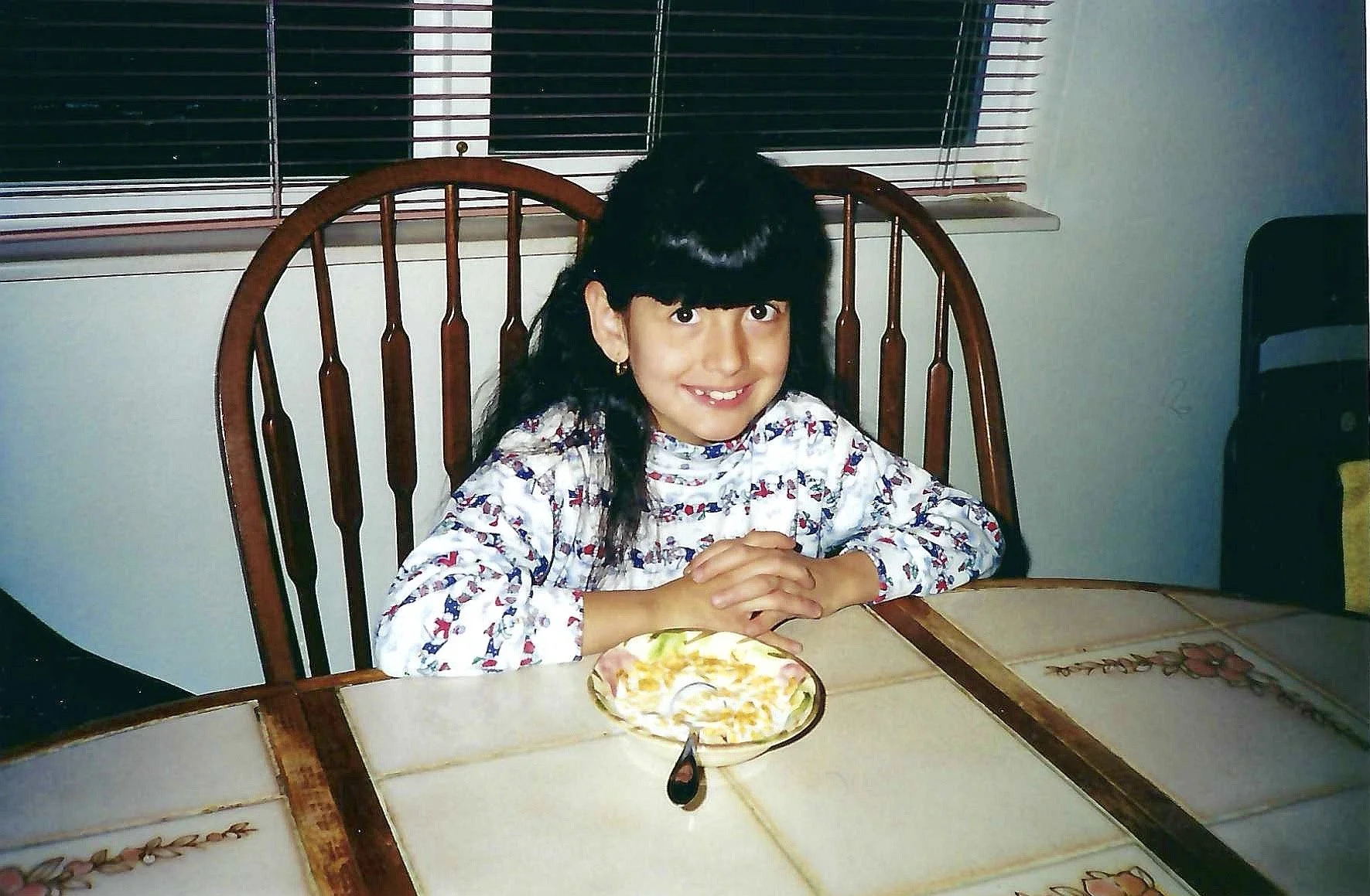 A young girl with black hair and bangs, wearing a long-sleeved shirt with red, blue, and white patterns, sitting at a kitchen table with a pizza on a plate. She is smiling with her hands clasped together on the table.