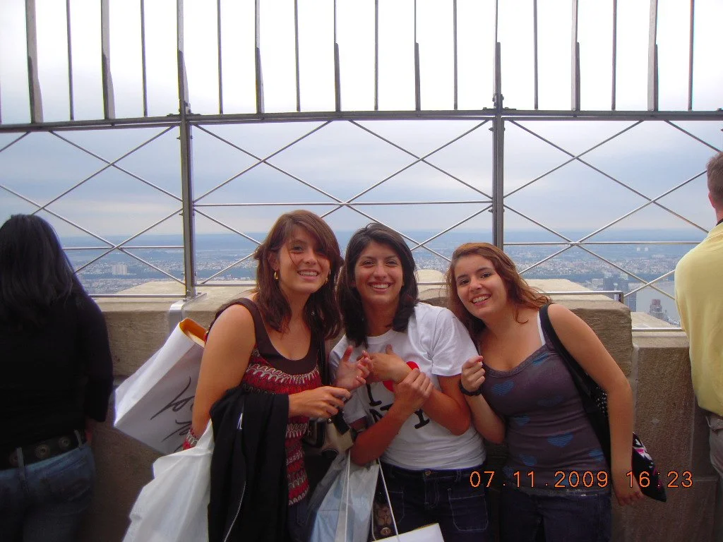 Three young women smiling at the camera on a rooftop observation deck with a cityscape view behind them, during daytime on November 7, 2009.