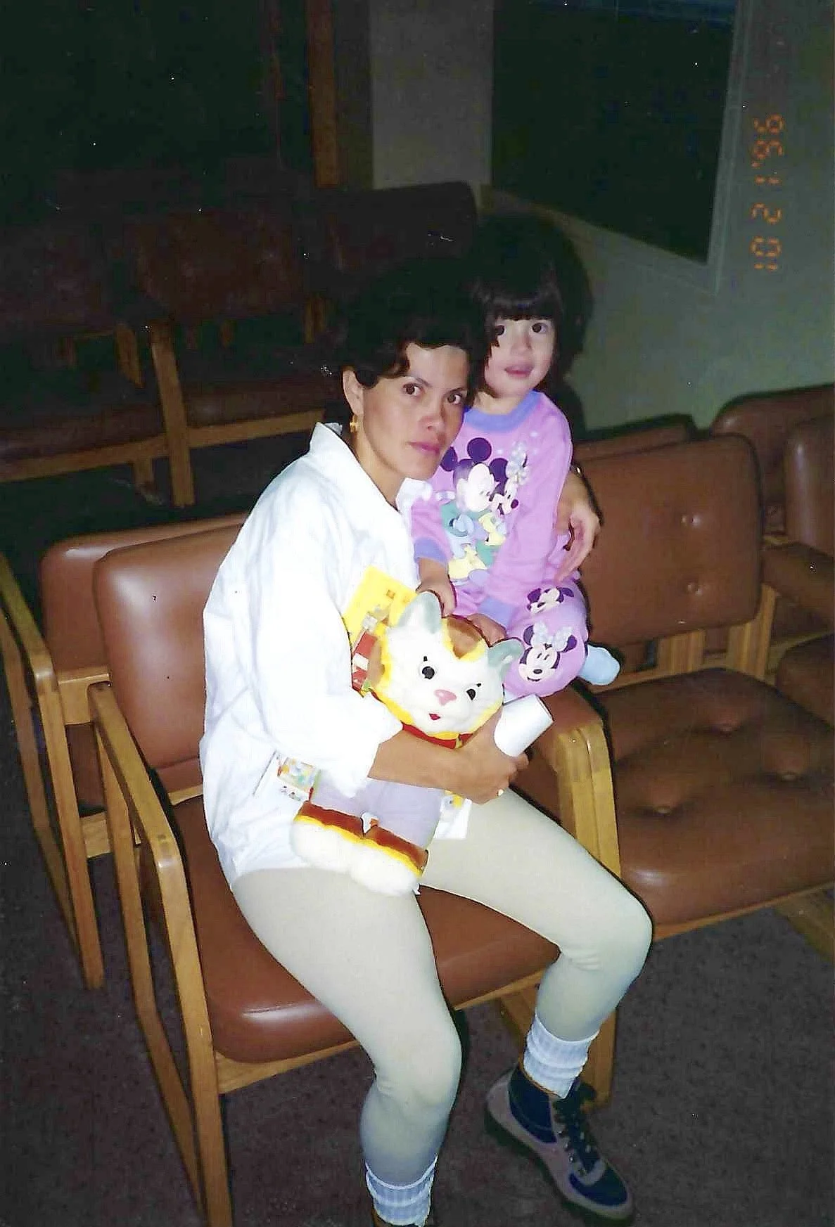 A woman and a young girl sitting on a brown leather bench in a dimly lit room. The woman is holding a plush toy cat, and both are looking at the camera. The woman wears a white shirt and light-colored pants, with socks and sneakers. The girl is in pi