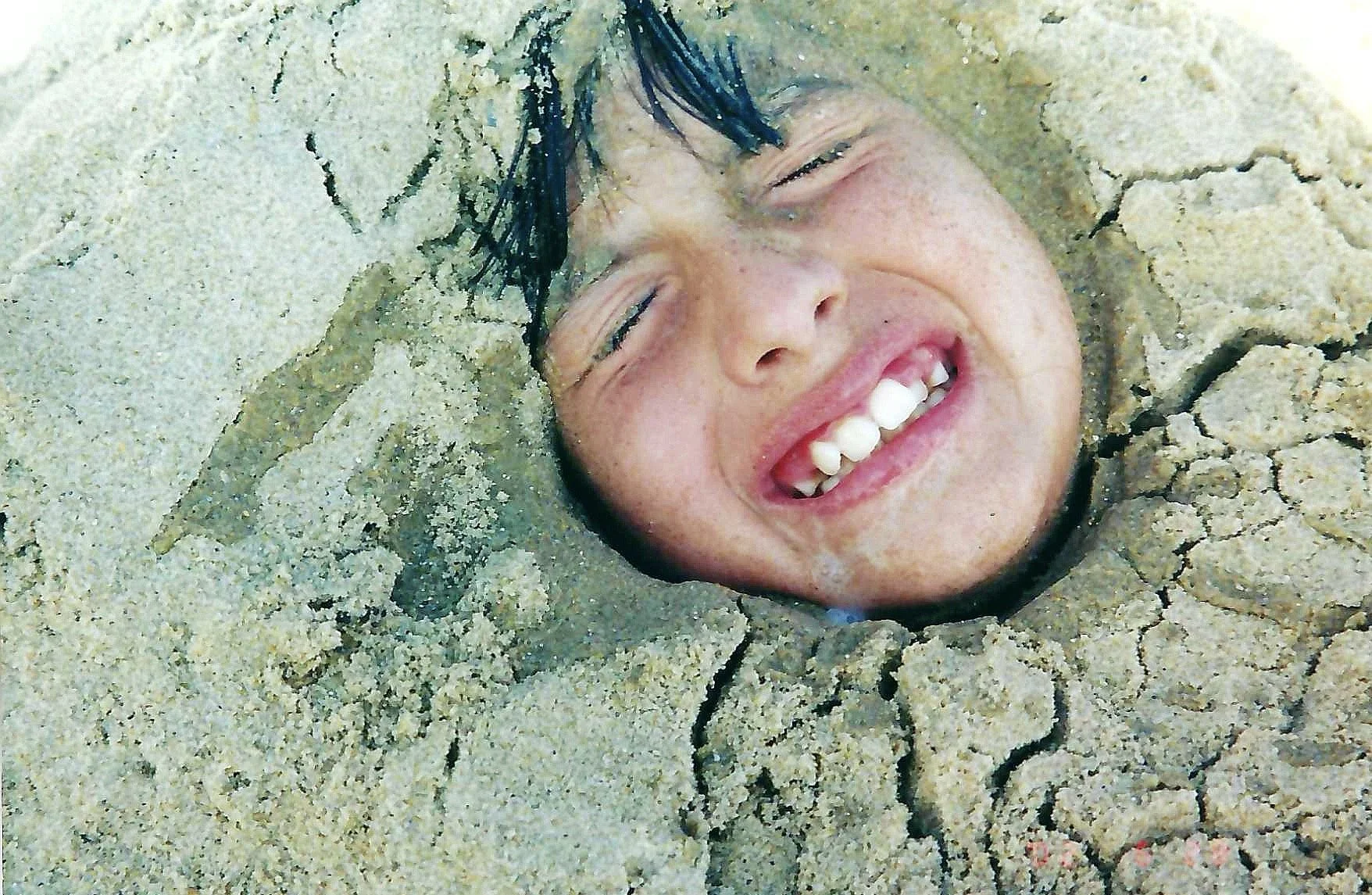 A person with black hair smiling and showing white teeth while their head is buried in the sand.