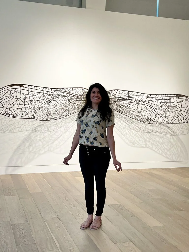 A woman smiling inside an art gallery, standing in front of a wall with a large sculpture resembling dragonfly wings.