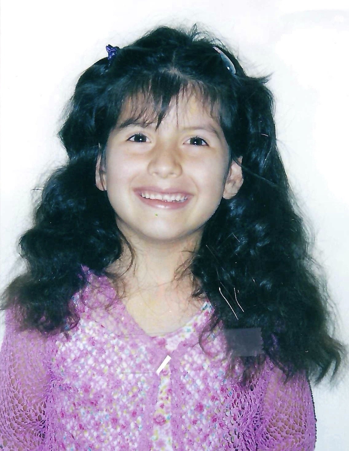A young girl with long, curly dark hair, smiling, wearing a pink top with a floral pattern.