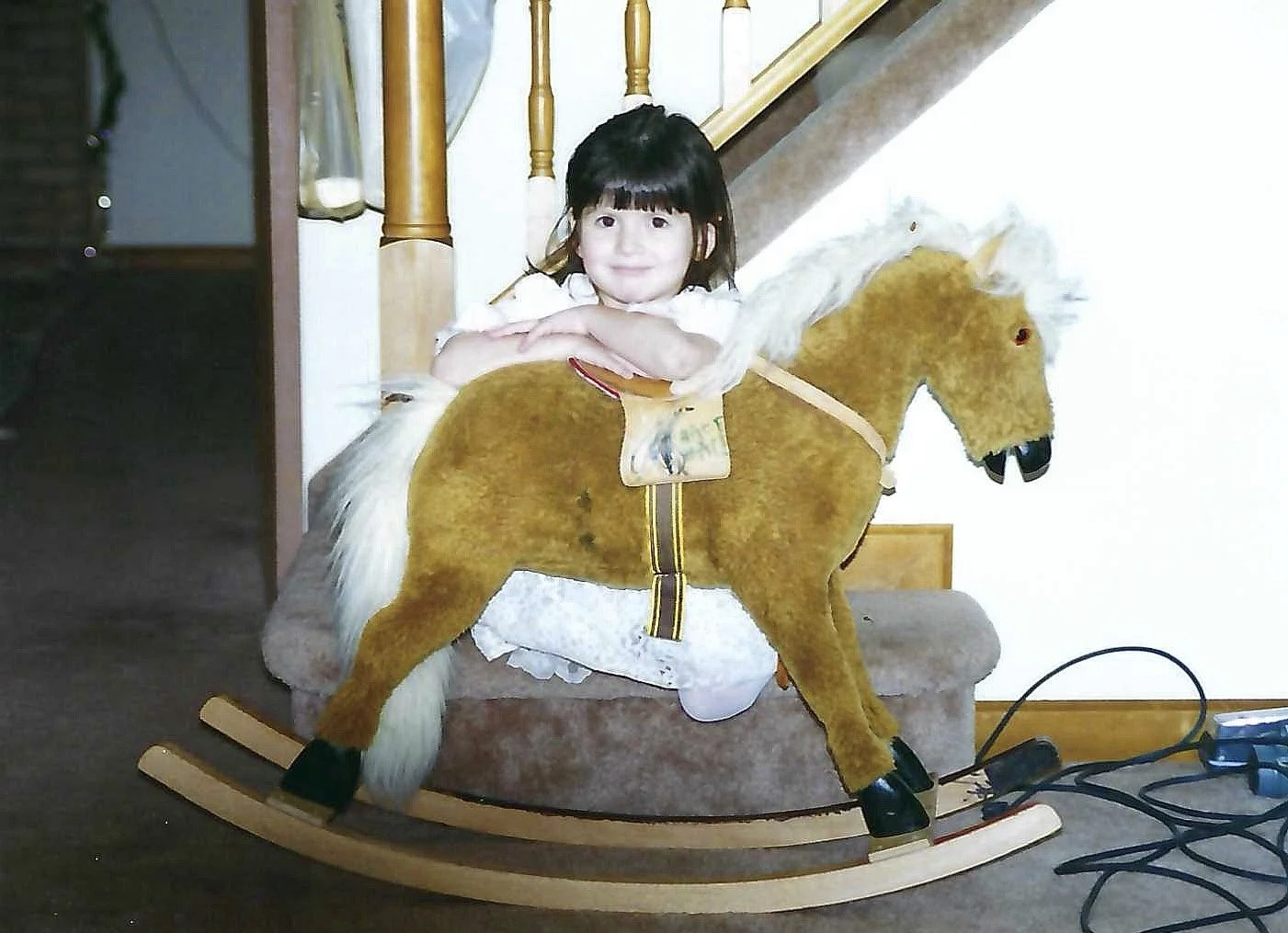 A young girl sitting on a rocking horse in a room with wooden furniture and a staircase nearby.