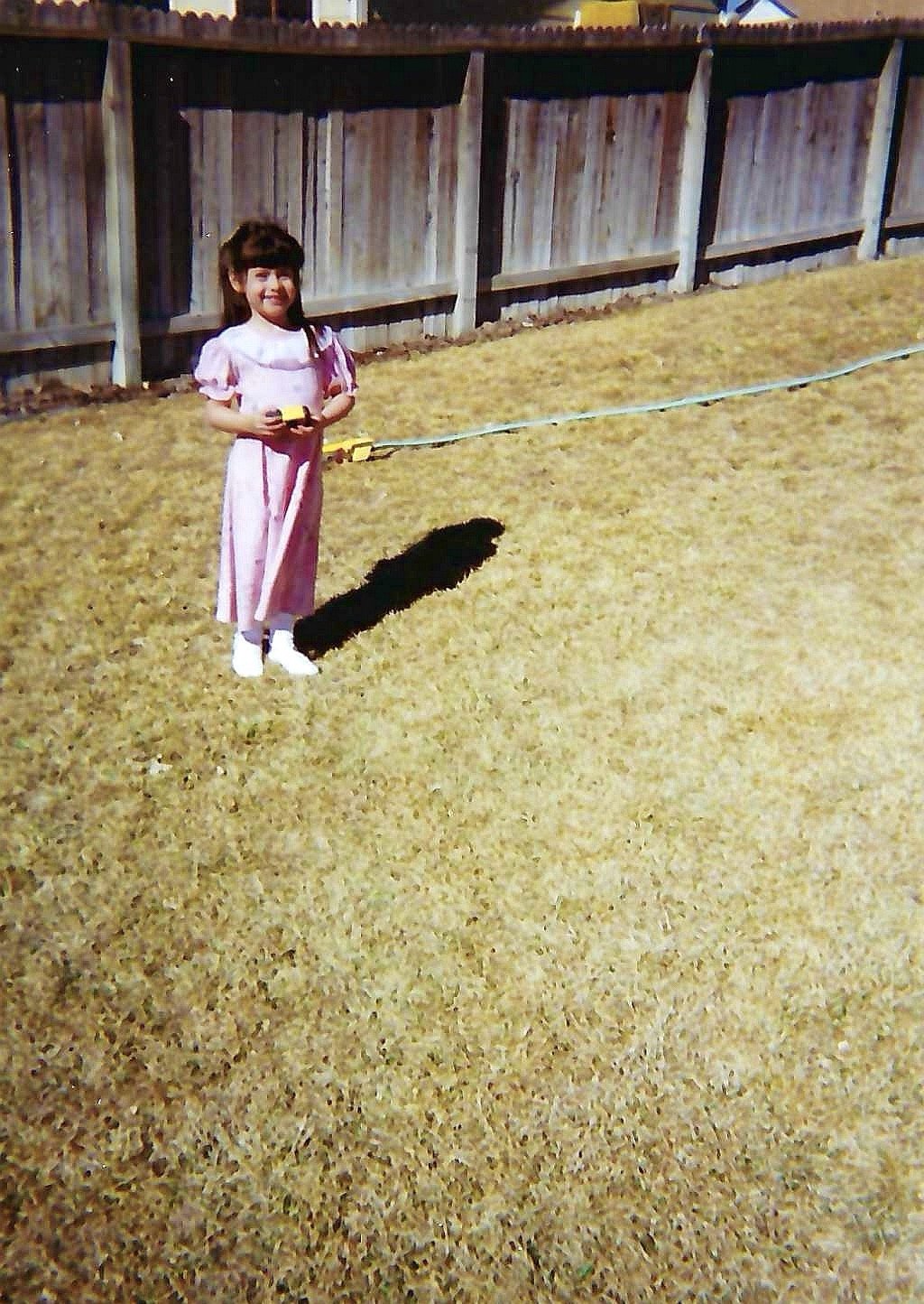 A young girl wearing a pink dress and white shoes in a backyard, holding a yellow toy and standing on a yellow lawn, with a wooden fence in the background.