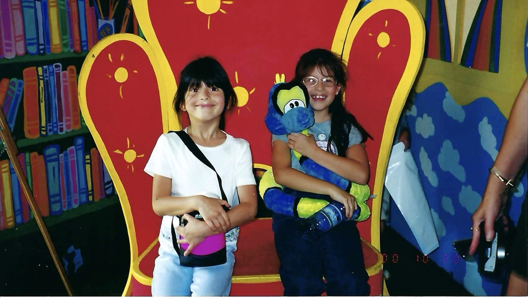 Two young girls smiling and posing in front of a colorful, cartoon-style backdrop. One girl has black hair, is wearing a white shirt and carrying a small bag. The other girl has glasses, is holding a plush toy, and has a water bottle. The backdrop fe