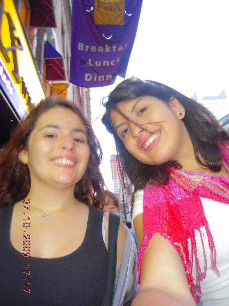 Two young women smiling for a selfie on a busy city street on a sunny day.