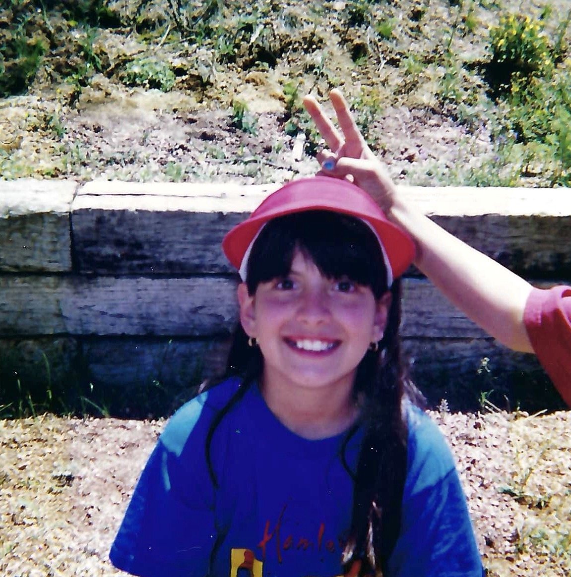 Girl with long dark hair wearing a blue t-shirt, smiling, and a red hat perched on her head, with a hand making a peace sign above her head, standing in front of a wooden fence and grassy, rocky background.