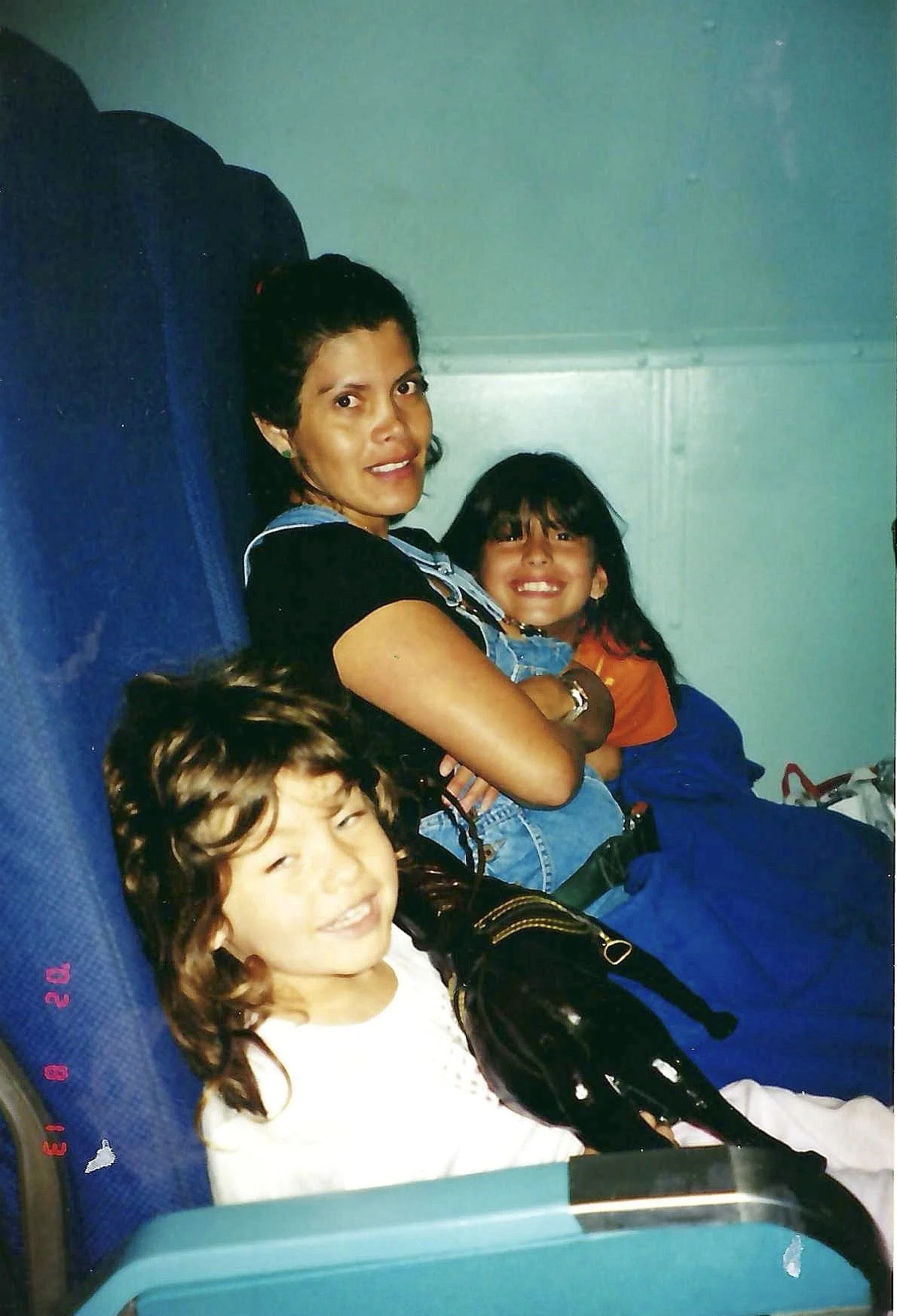 A woman and two children sitting on airplane seats, smiling at the camera. The woman is holding a young girl, and a boy with curly hair is sitting in front, partially visible.