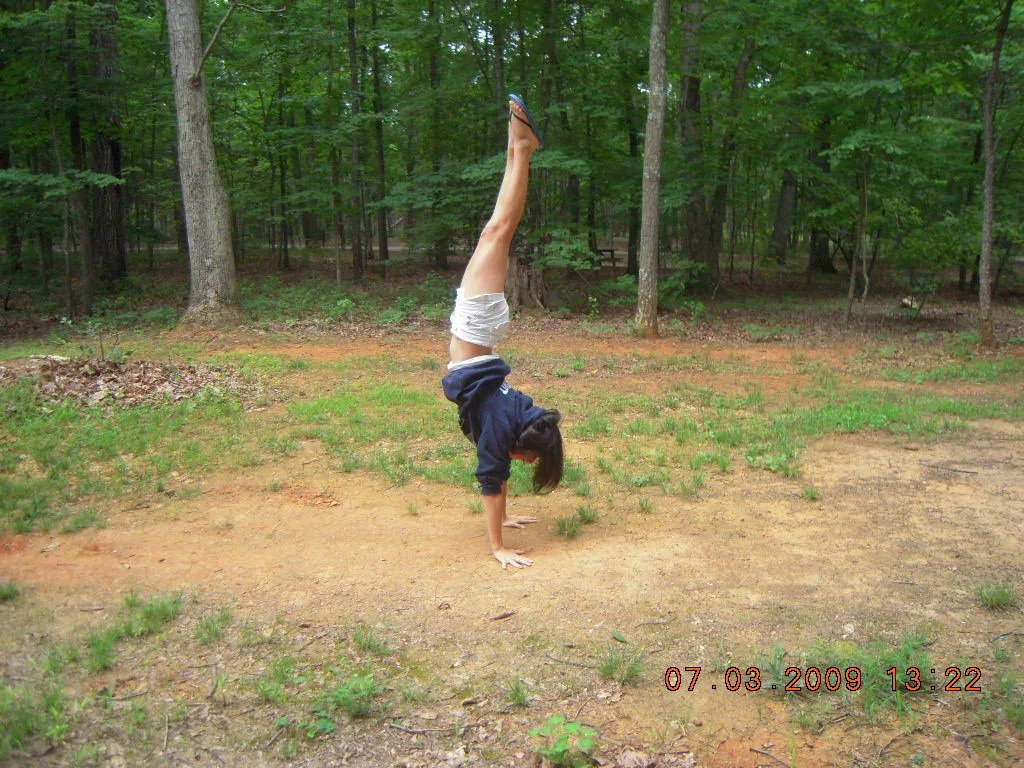 A girl doing a handstand in a forested area with green trees and a dirt ground.