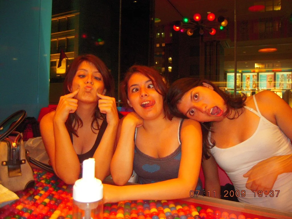 Three young women making funny faces at a table in a colorful restaurant or bar, with a multicolored tablecloth, a purse, and a bottle of hand sanitizer visible in the foreground.