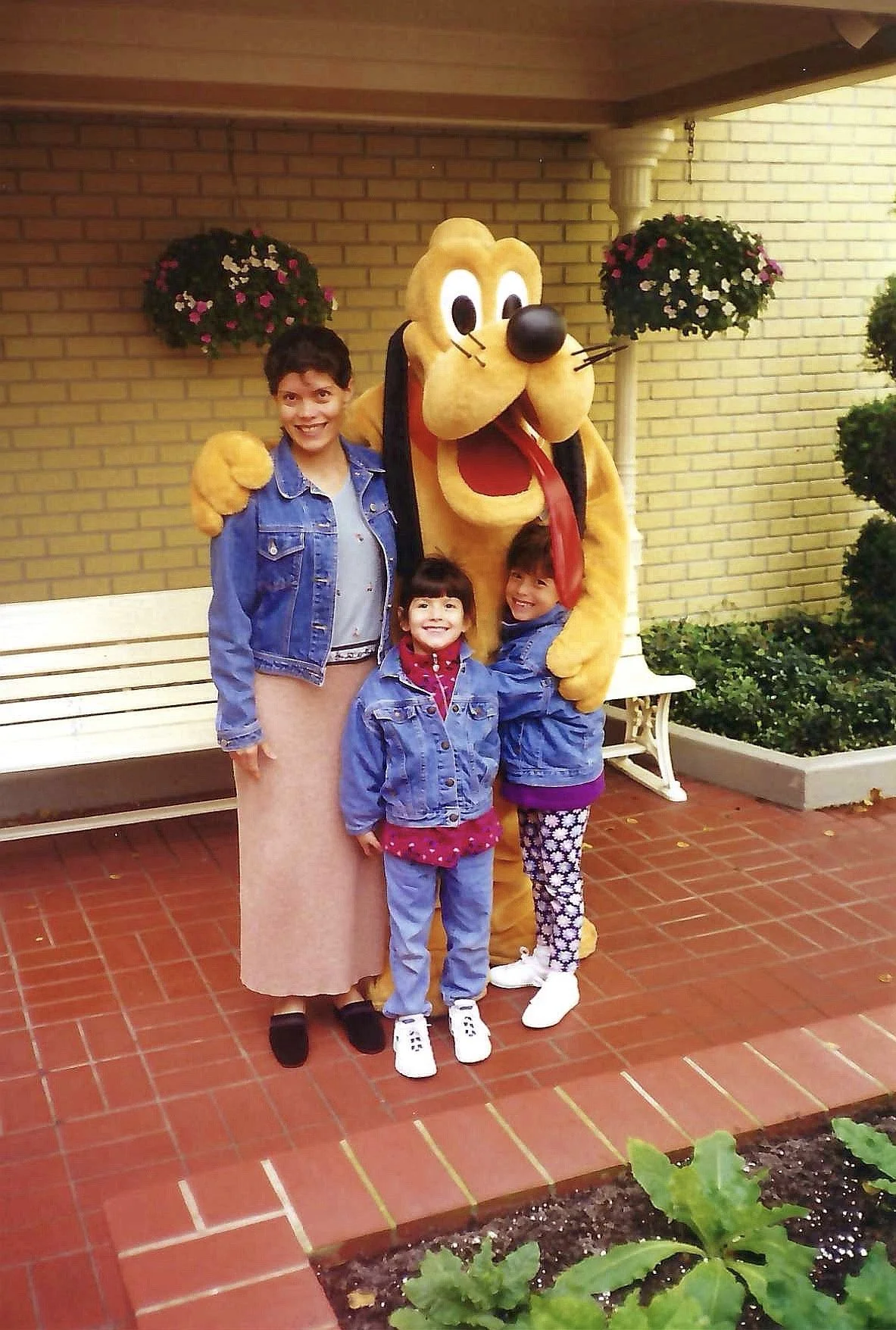 Three children and a person in a Pluto costume pose for a photo outdoors, with brick and yellow walls, potted flowers, and a bench in the background.