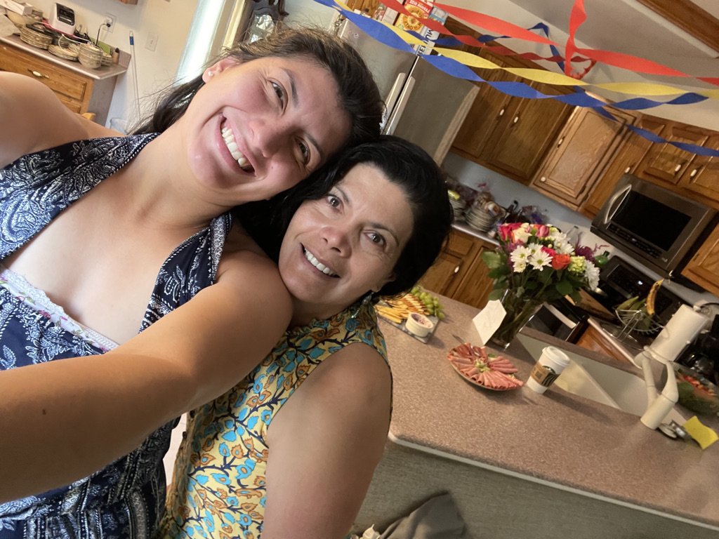 Two women smiling and taking a selfie in a kitchen decorated for a celebration, with colorful streamers hanging from the ceiling and a bouquet of flowers on the counter.