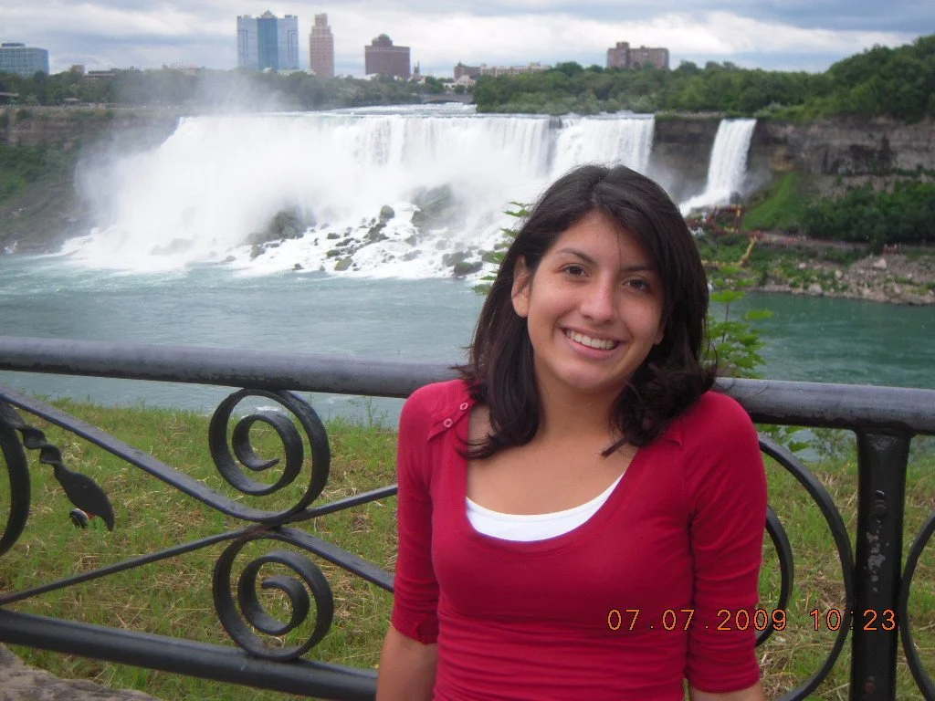 Young woman with dark hair smiling at the camera in front of Niagara Falls with city skyline in the background.