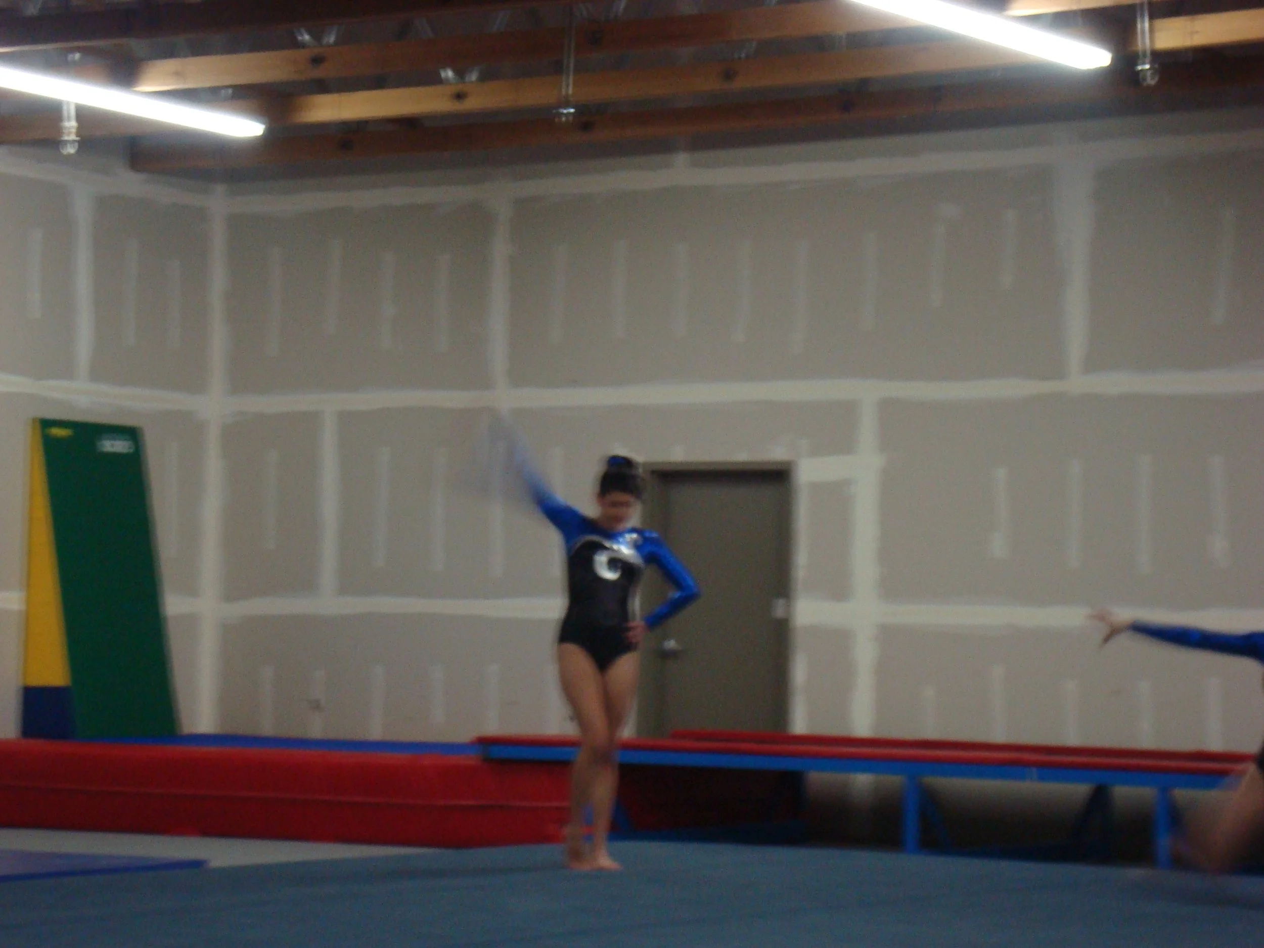 A gymnast in a blue and black uniform standing on a trampoline in an indoor gym, with gym mats and a person in the background.