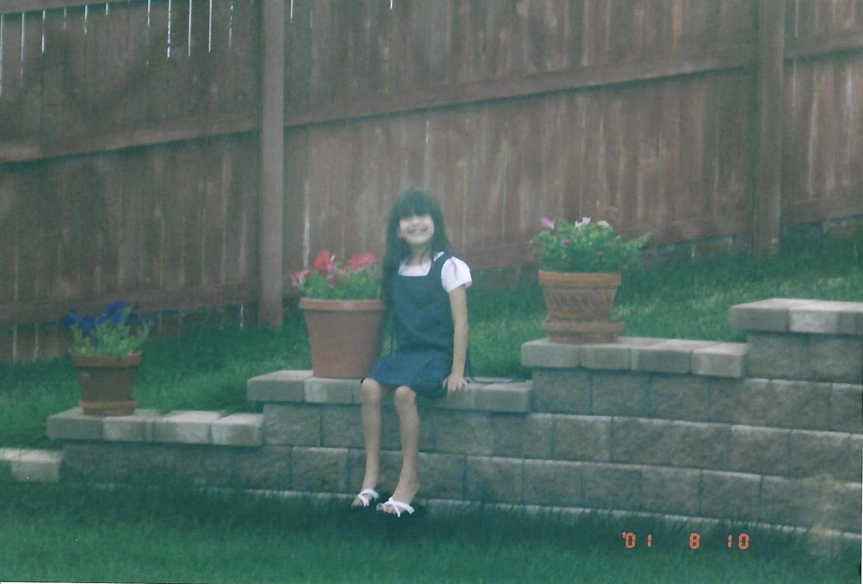 A young girl with long dark hair and a white face mask, wearing a navy blue dress with white shirt sleeves underneath, sitting on a step in a garden area. She is surrounded by potted flowers and a wooden fence.