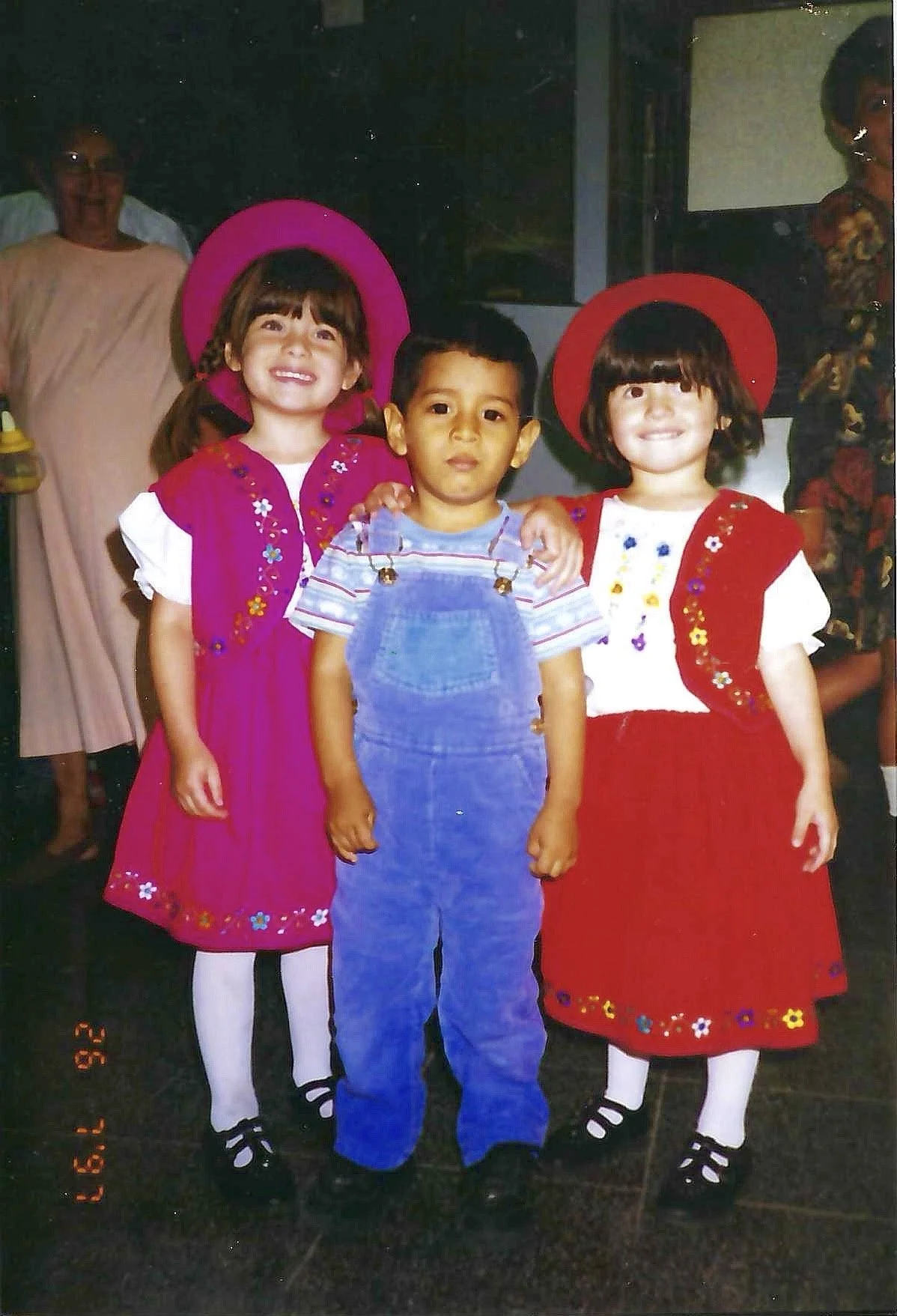 Three children standing together, two girls wearing red and pink dresses with wide-brimmed hats, and a boy in overalls and a striped shirt, smiling and posing for a photo indoors.