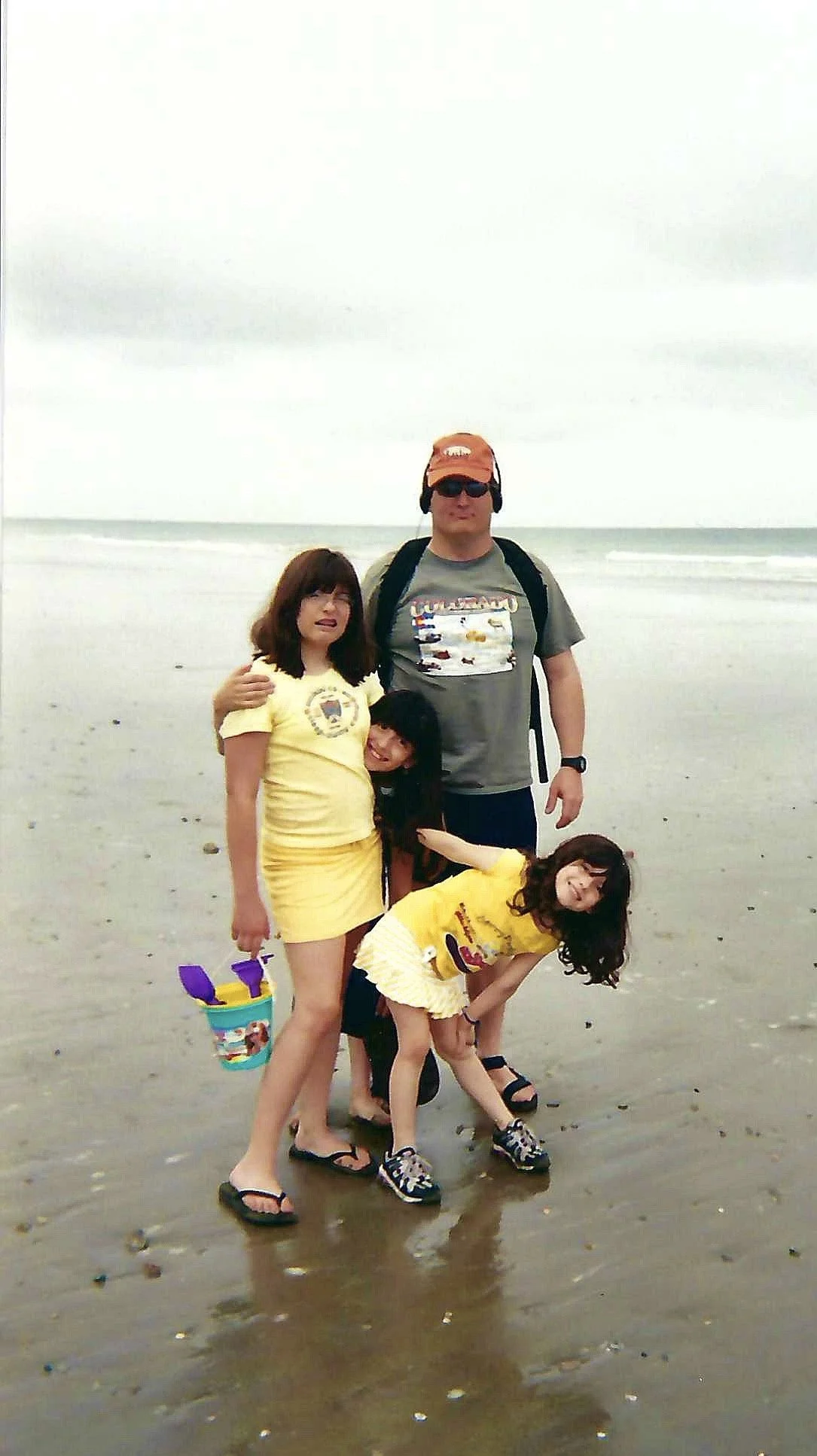A family of five on the beach with a cloudy sky, including a man with a backpack, sunglasses, and cap, a woman in a yellow outfit, and three young girls in yellow dresses, one holding a bucket, all smiling and posing for the photo.