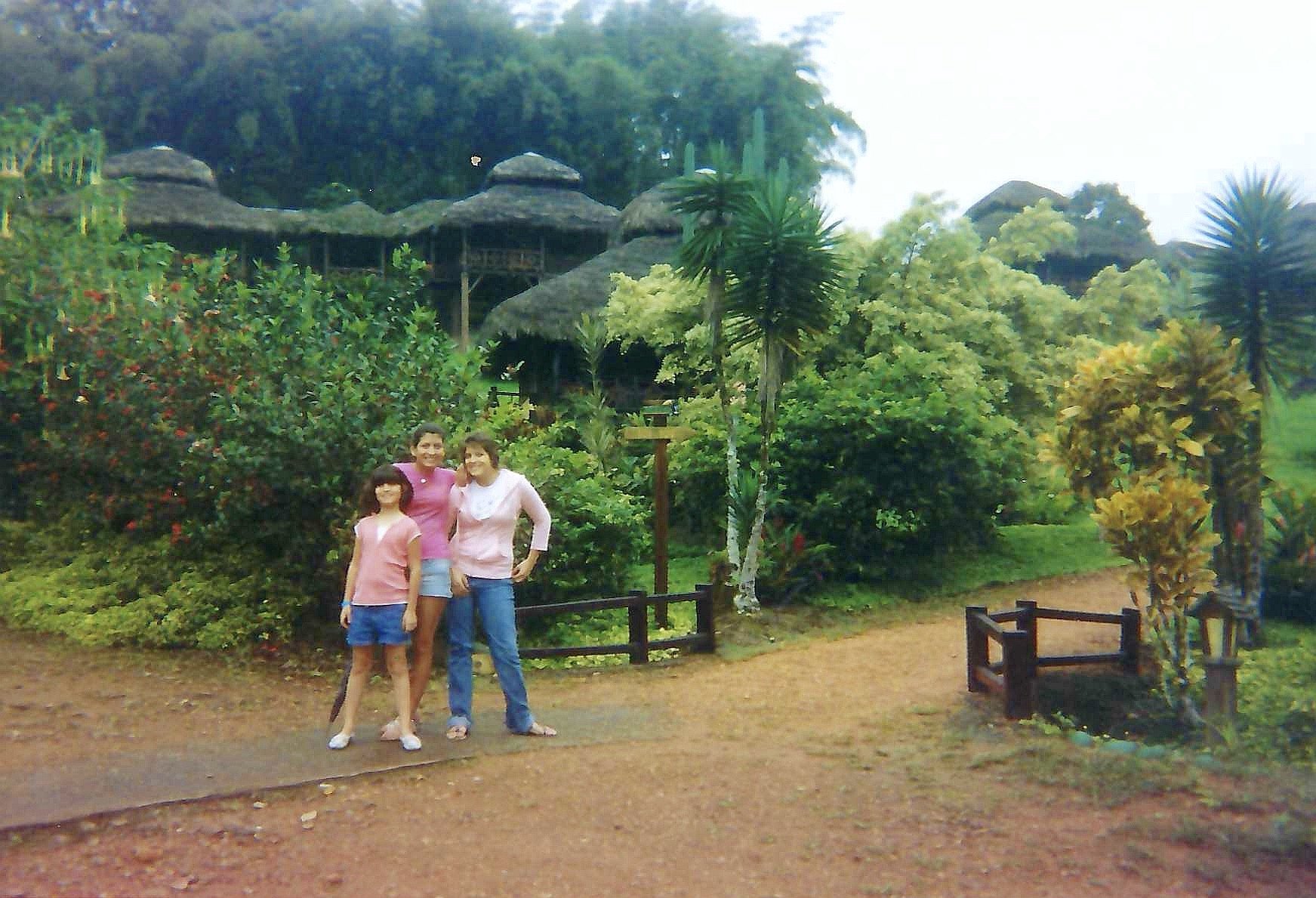 Three women and a girl standing on a dirt path in a lush, green garden with tropical plants and trees, and thatched-roof structures in the background.