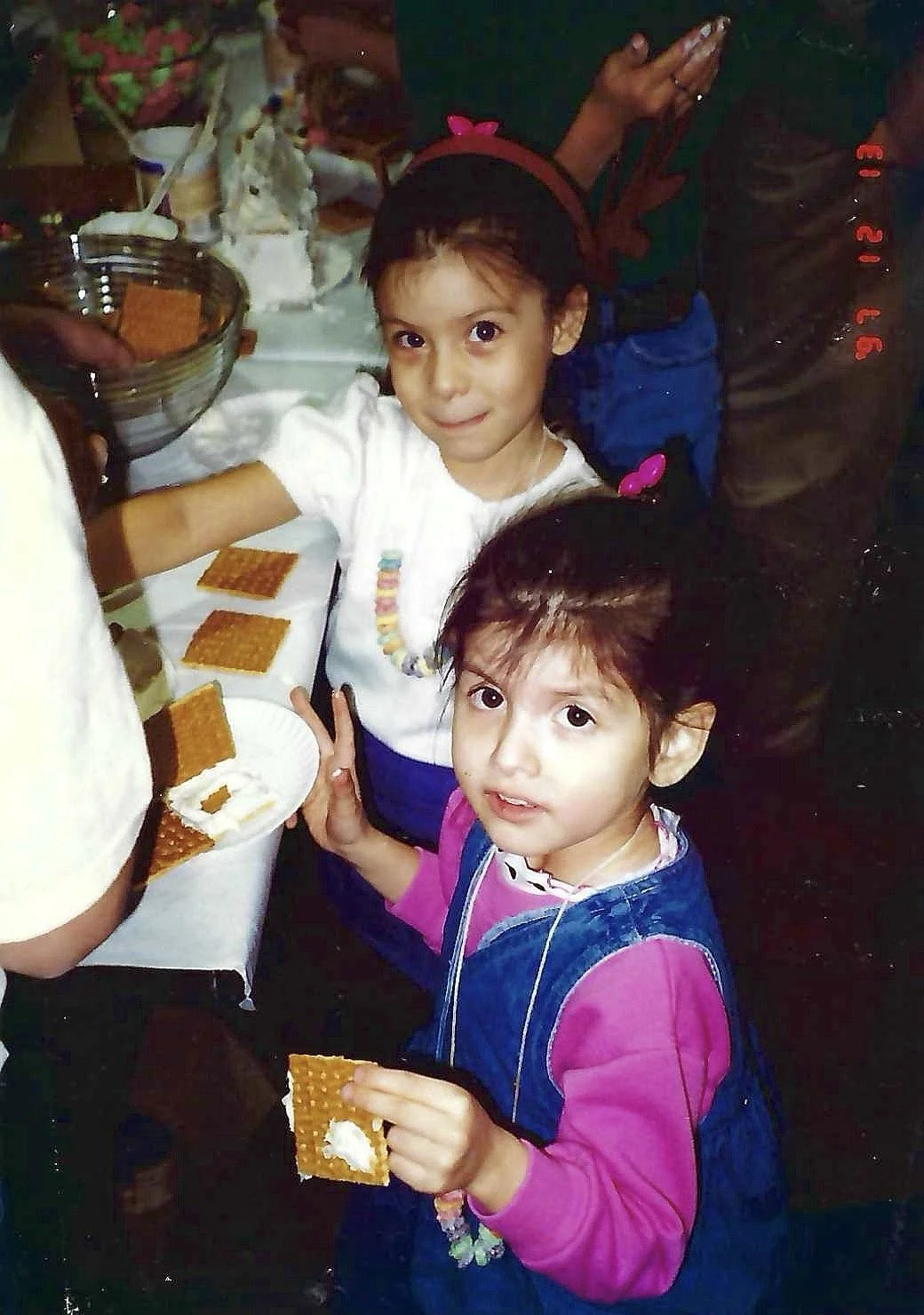 Two young girls at a table filling graham crackers with cream for a s'mores snack. One girl wears a white shirt and a colorful necklace, while the other wears a pink and blue jacket. There are other people in the background preparing food. The scene 