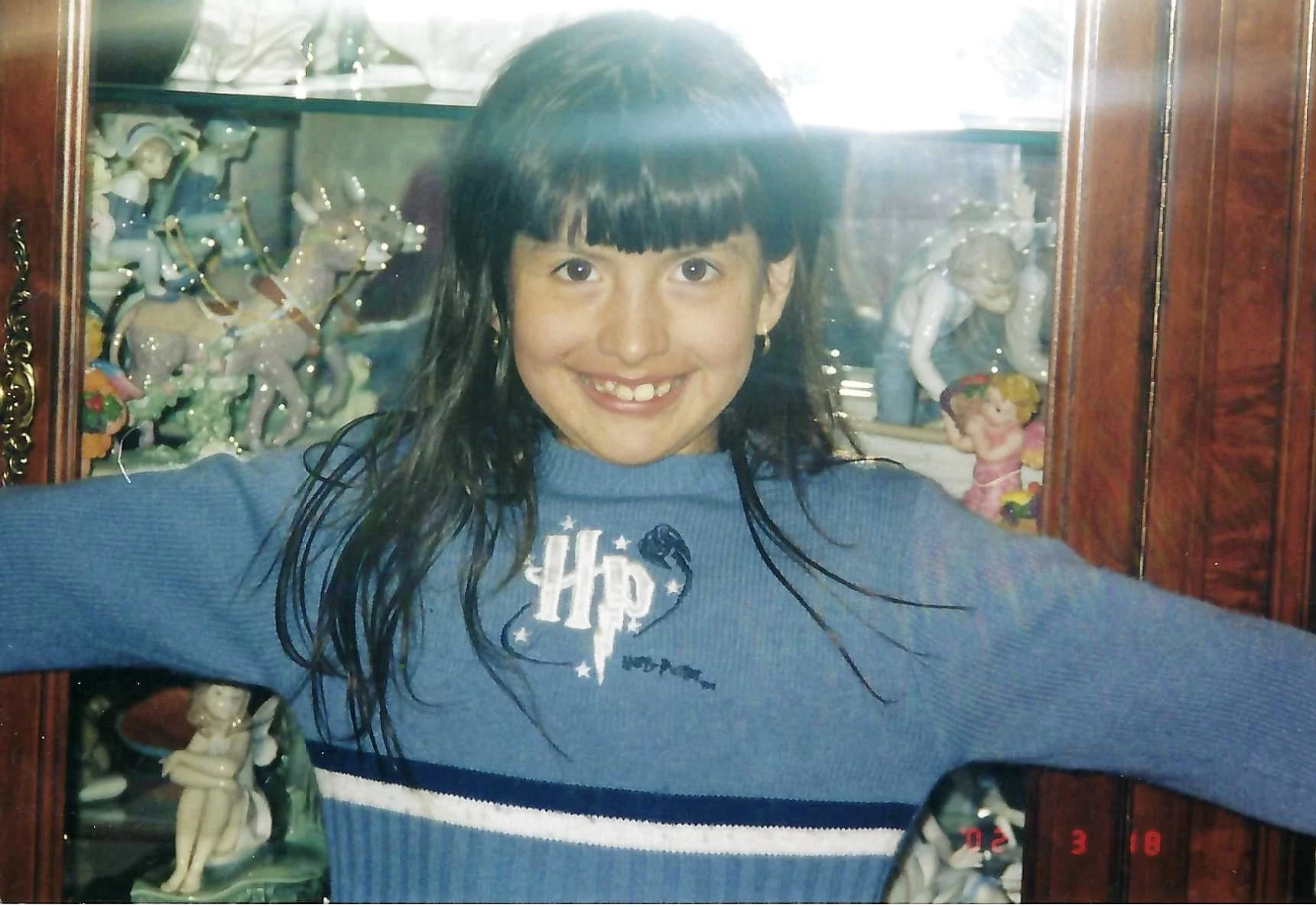 A girl with dark wet hair and a bright smile taking a selfie in front of a display cabinet filled with porcelain figurines and decorations.