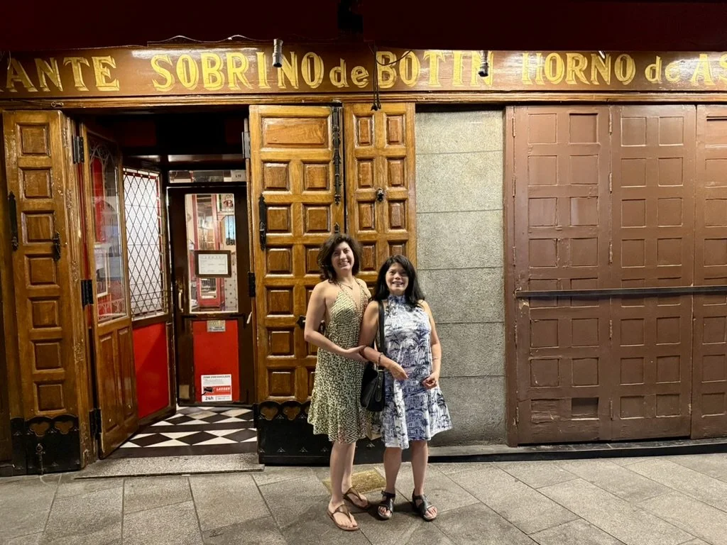Two women standing outside a restaurant with a wooden facade and a sign above it. Both women are smiling and wearing summer dresses.