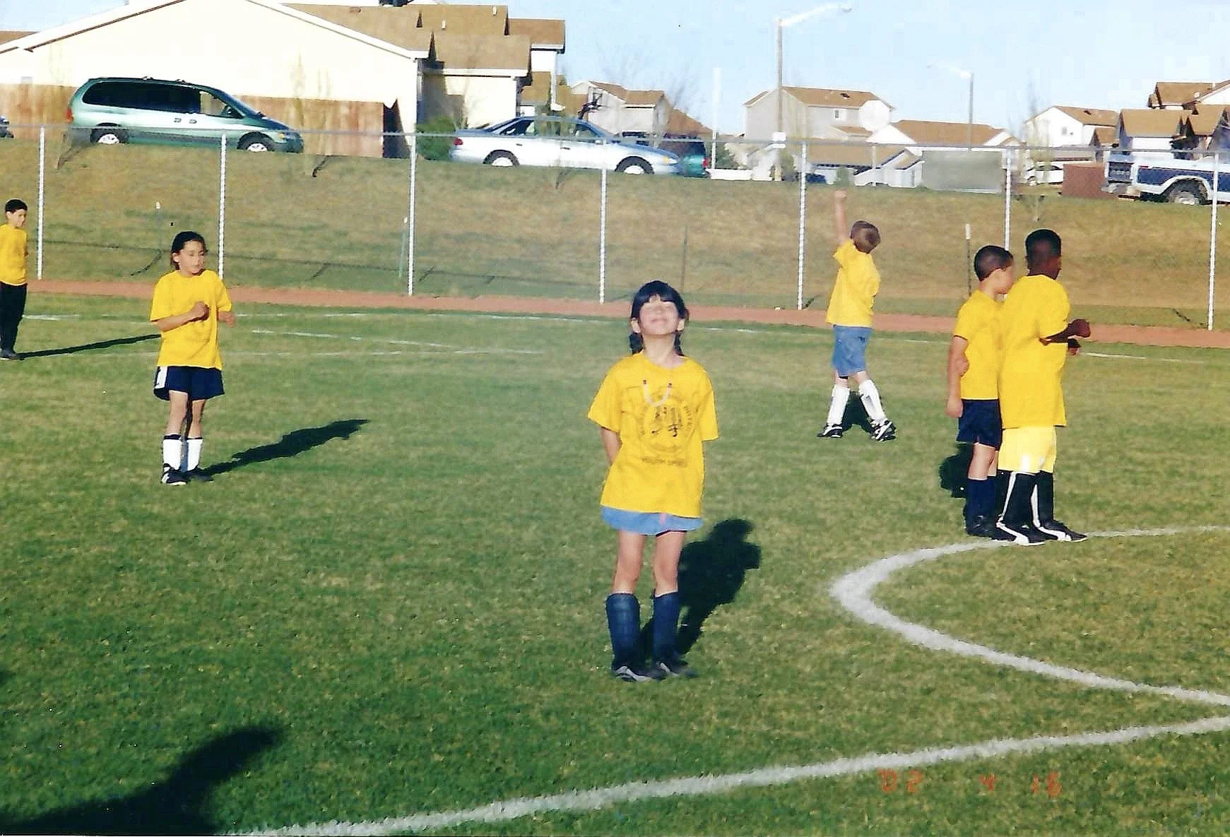 Children playing soccer on a field, wearing yellow jerseys and black shorts, with houses and cars in the background.