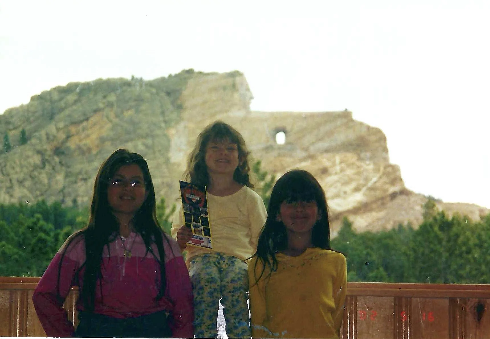 Three young girls standing in front of the ancient ruins of the Colosseum, with greenery in the foreground and a bright sky overhead.