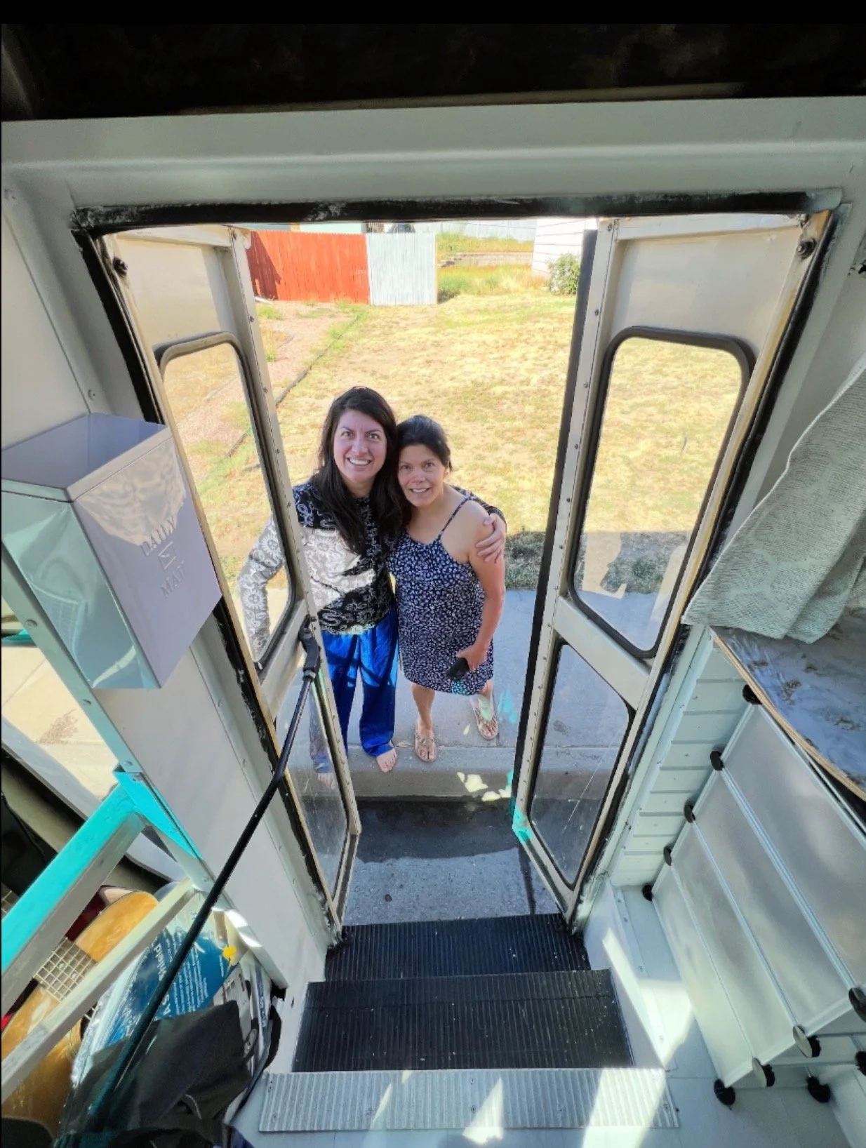 Two women standing outside a food truck, smiling at the camera, one wearing a patterned dress and the other in a top with blue pants, holding a phone, with a grassy yard and a red and gray fence in the background.
