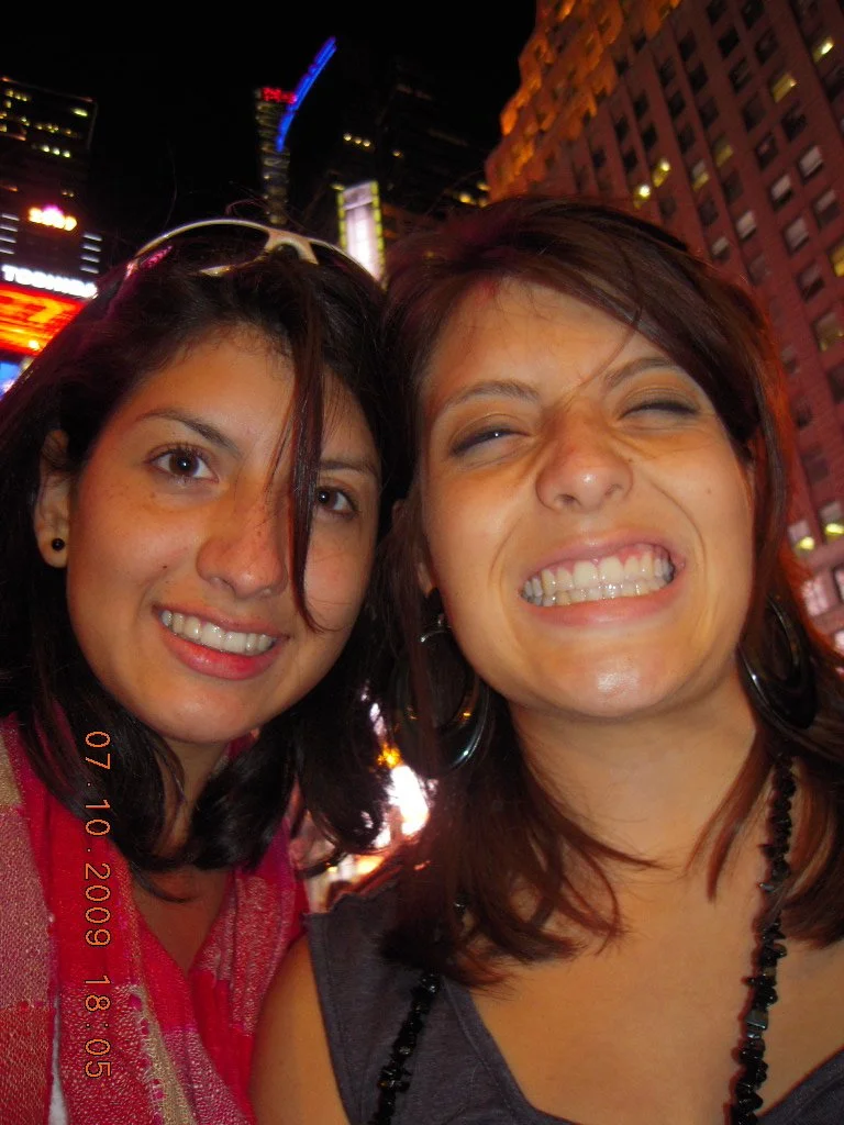 Two women smiling and taking a selfie at night in an urban area with bright city lights and tall buildings in the background.