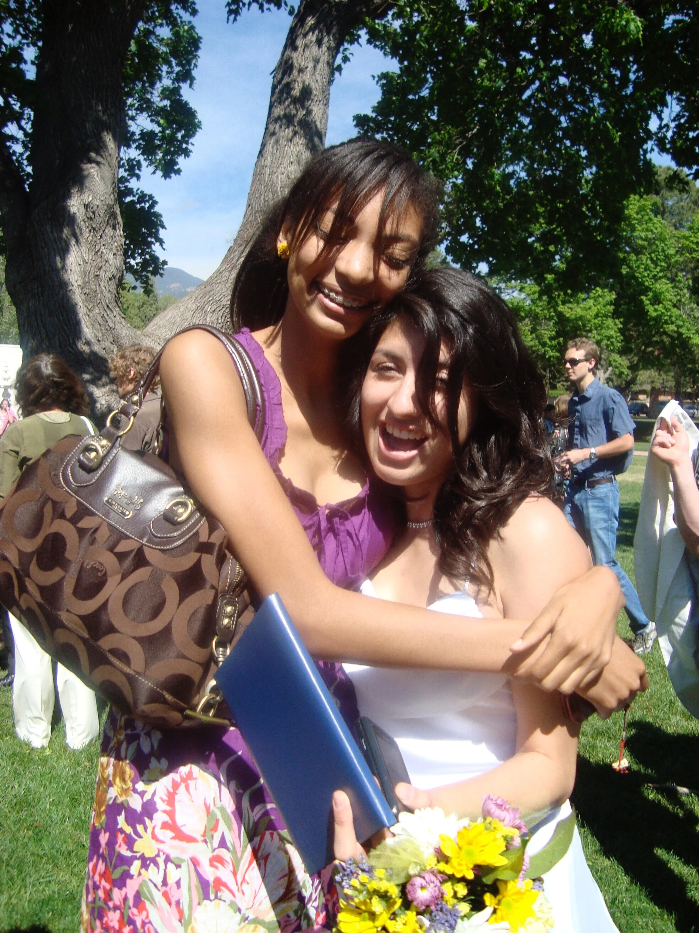 Two women embracing, smiling, outdoors under a tree, with students and mountains in the background. One woman holds a bouquet of flowers, and the other holds a blue folder.