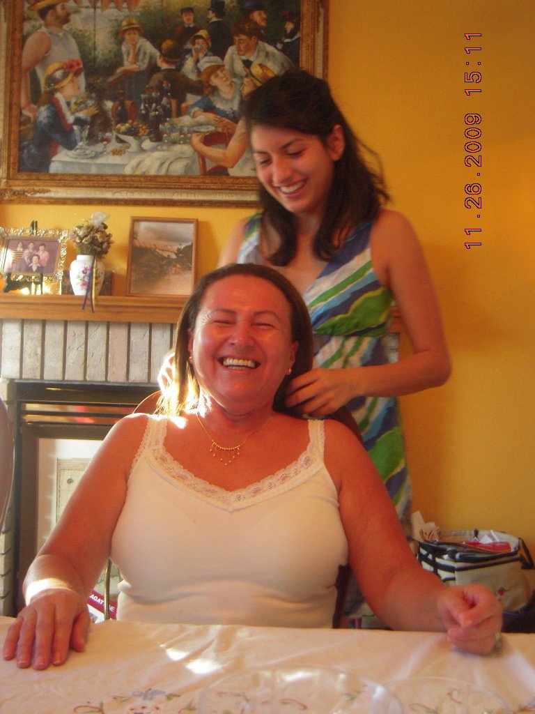 A woman sitting at a table smiling, with a young girl standing behind her, also smiling, in a cozy room with a painting and framed photos on a mantelpiece.