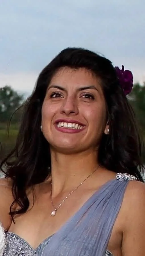 A woman with dark hair, smiling, outdoors with a cloudy sky, wearing a purple dress with beaded shoulder straps, a necklace with a pearl pendant, and a flower in her hair.