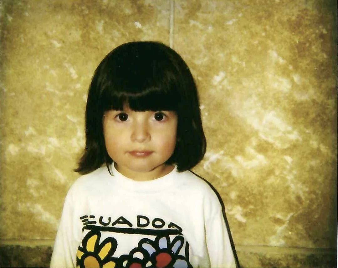 A young girl with black, shoulder-length hair and a straight, blunt cut bangs. She has light skin and is wearing a white t-shirt with colorful flower and text design. The background is a textured, yellowish-brown wall.
