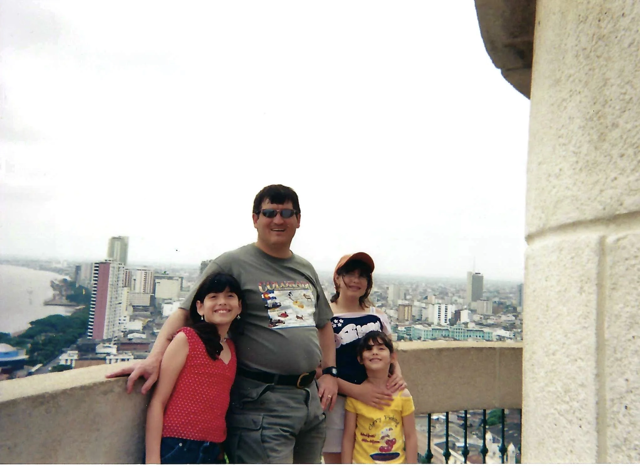 A group of people, including a man wearing sunglasses and four children, standing together on a balcony with a cityscape in the background.