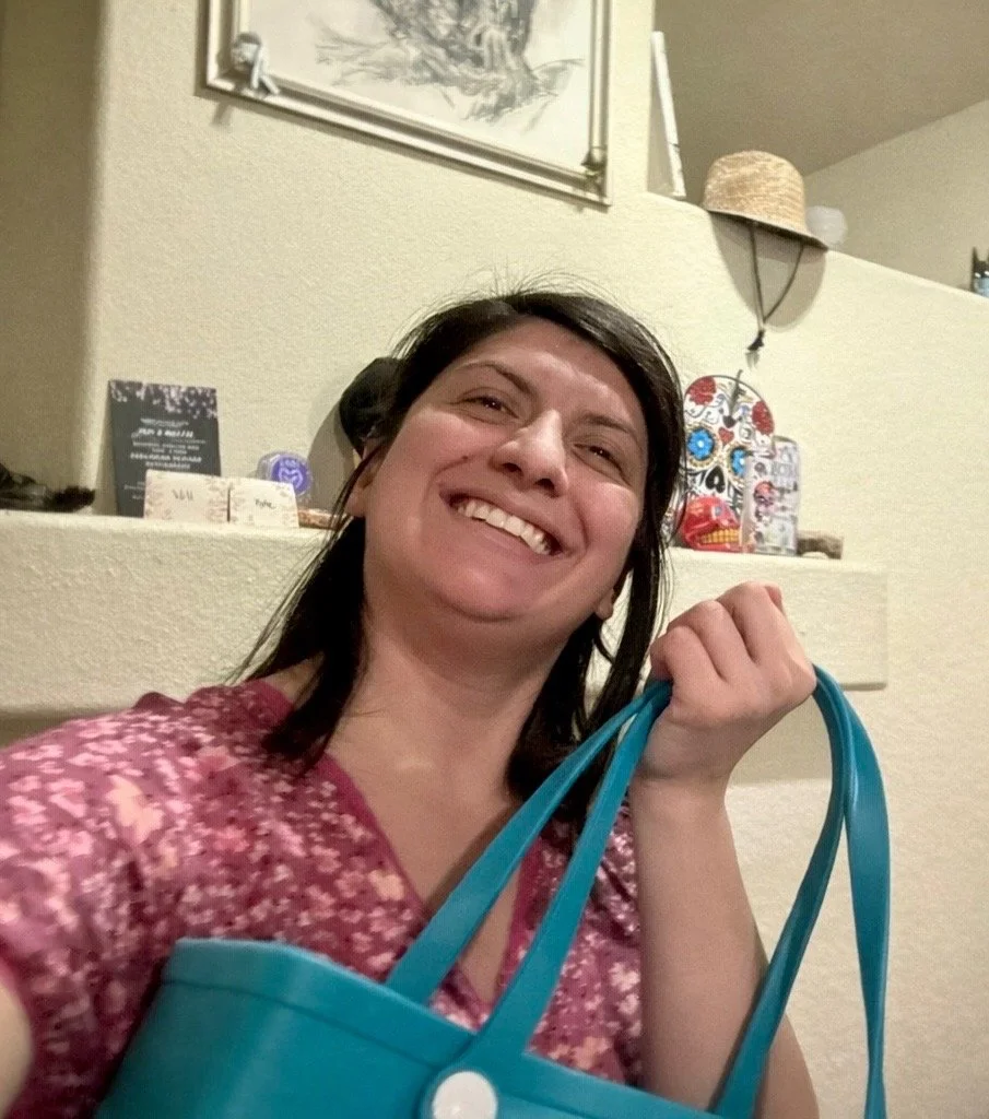 A woman with dark hair smiling and holding a blue reusable shopping bag, inside a home with a beige wall and decorative items in the background.