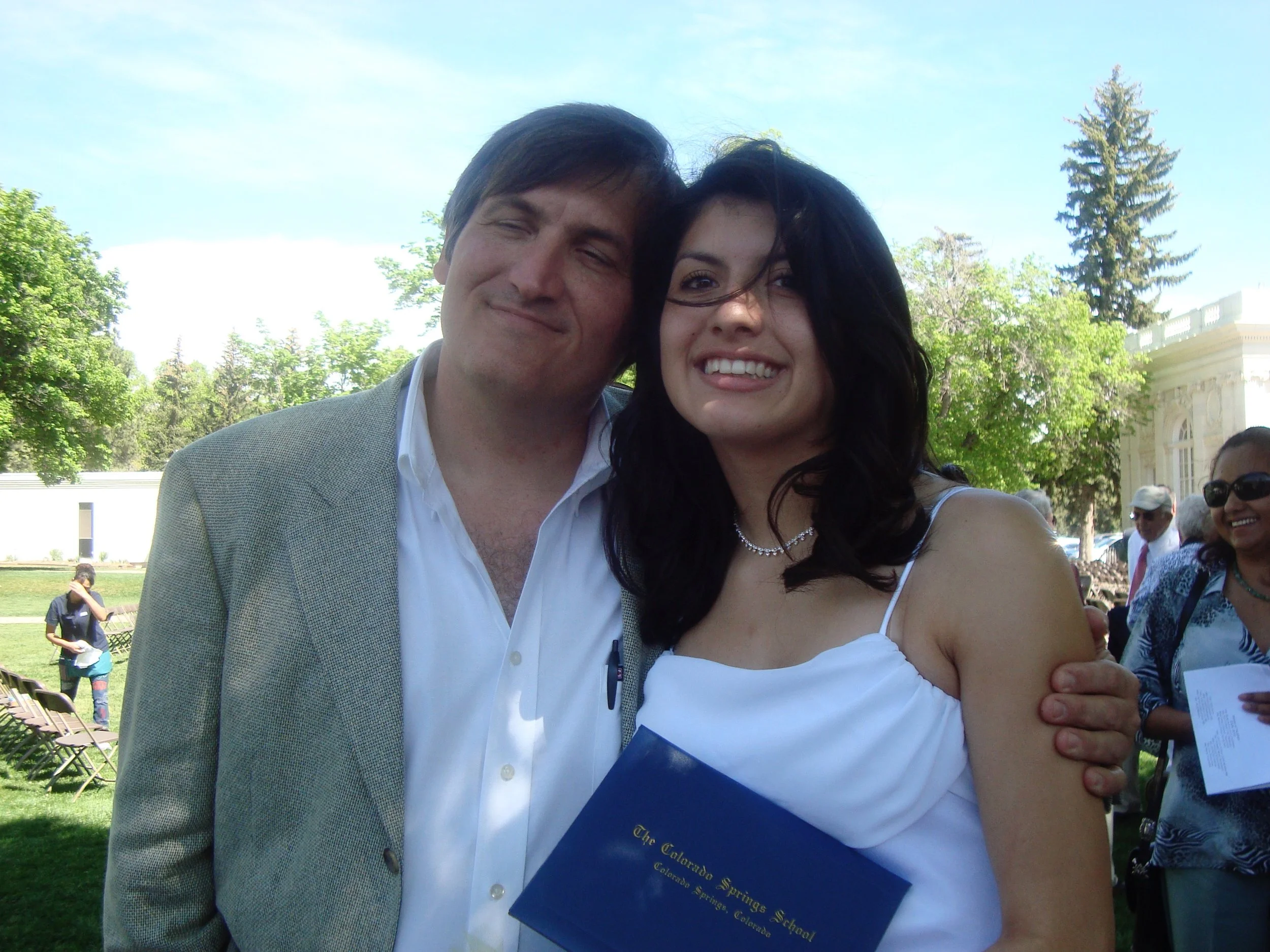 A man and woman hugging at an outdoor event, with trees and a building in the background, and a blue ribbon indicating a graduation or award.