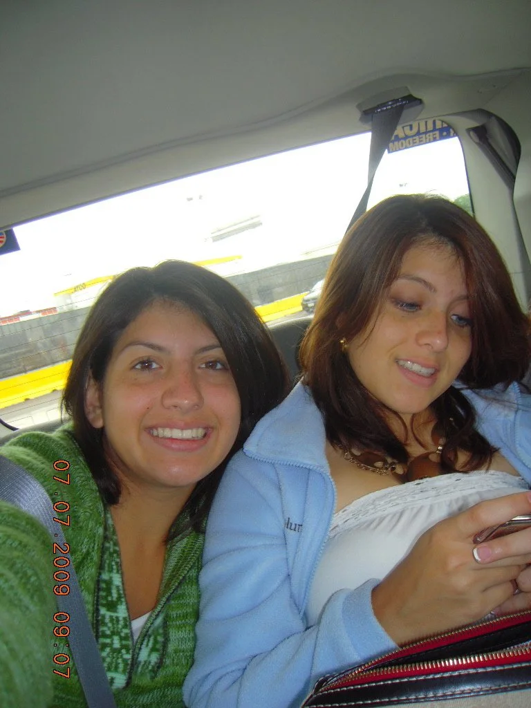 Two young women sitting in the backseat of a vehicle. One is smiling at the camera, and the other is looking at her phone. The background shows a highway with a guardrail and some vehicles.